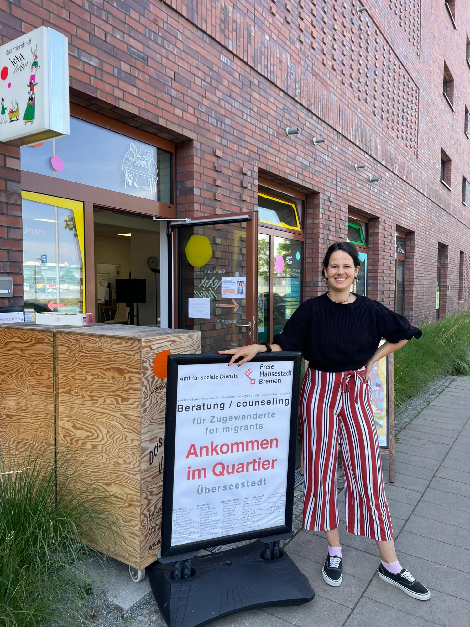 Kerstin Panzer steht an einer Werbetafel auf der "Ankommen im Quartier" geschrieben steht. Sie steht vor dem Quartierstreff Jetzt Hier. Einem Backsteingebäuden mit bunt beklebten Fenstern. Die Türe zum Quartierstreff ist geöffnet.