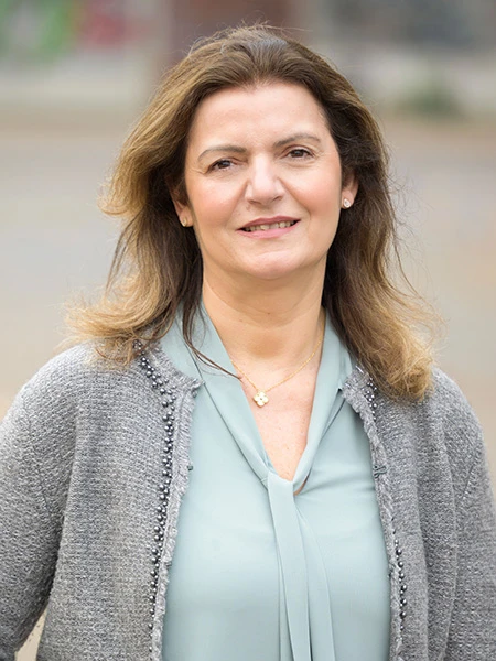 Frau mit schulterlangen, gewellten Haaren tr&auml;gt eine graue Strickjacke &uuml;ber einer hellblauen Bluse und eine goldene Halskette mit kleinem Anh&auml;nger