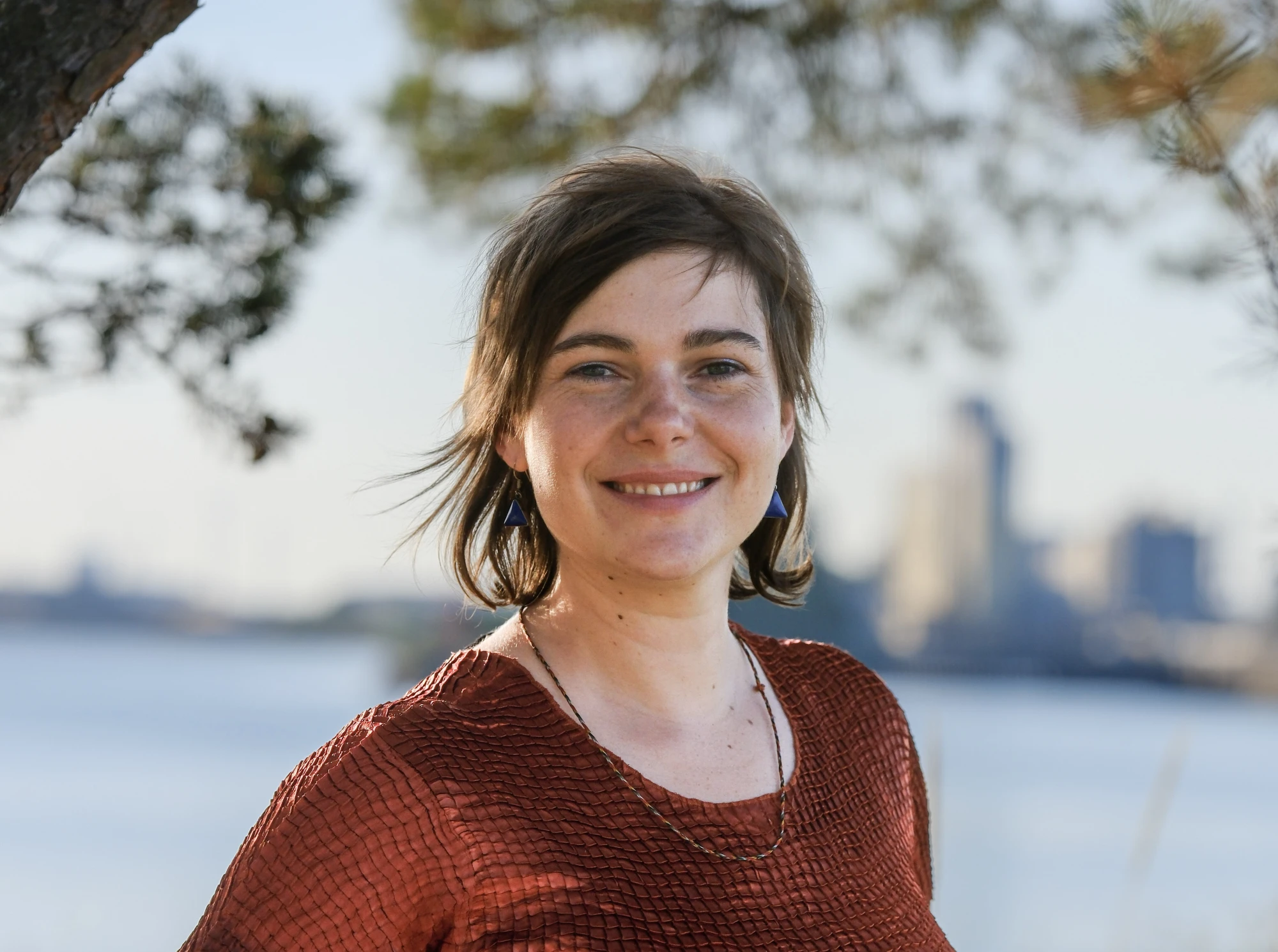 Frau mit mittellangen braunen Haaren steht in der Natur. Im Hintergrund sind Fabrikanlagen am Hafen zu erkennen.