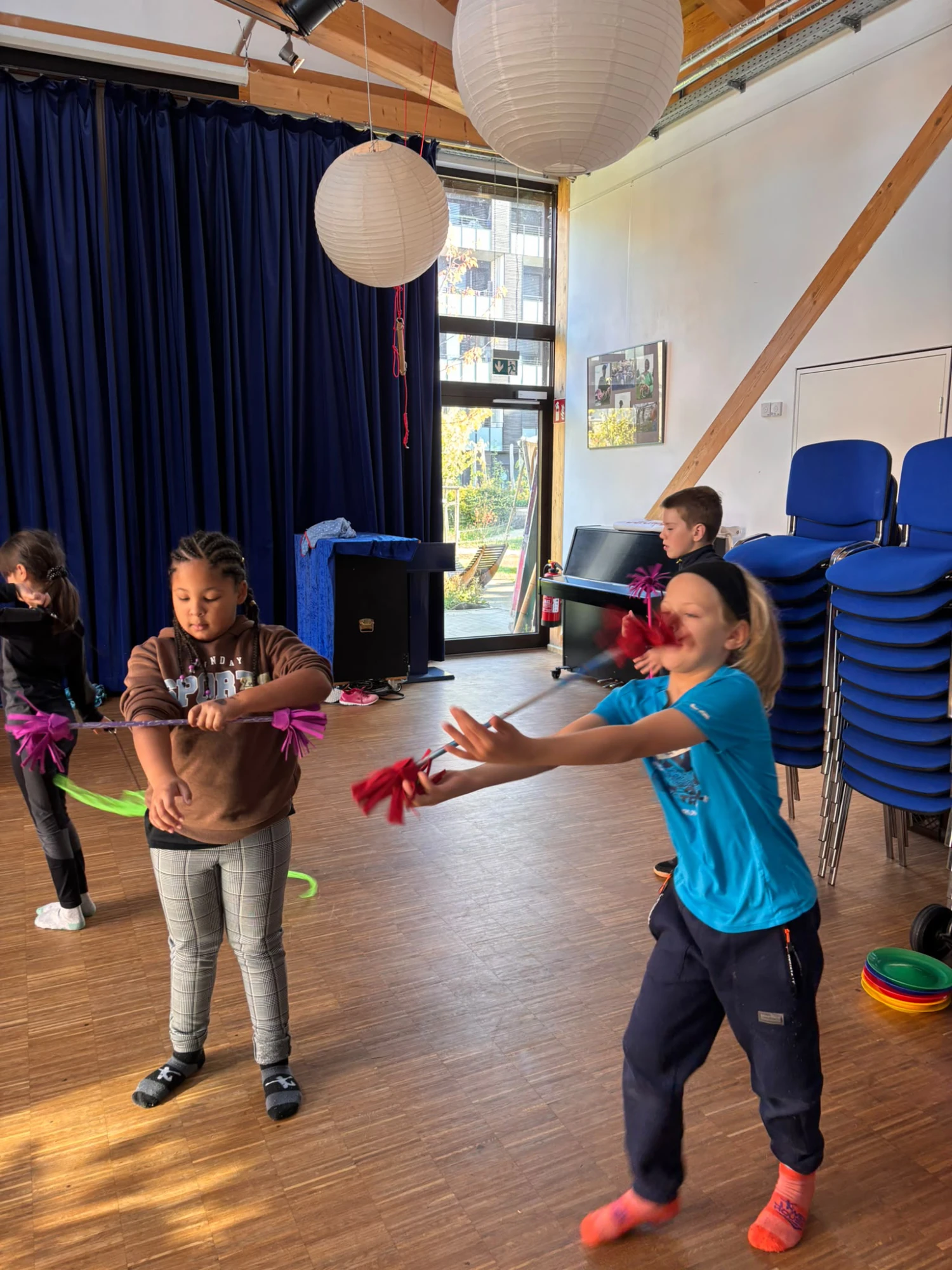 Kinder in einem Raum mit Holzboden und blauen Stühlen halten rote Pompons und bewegen sich spielerisch