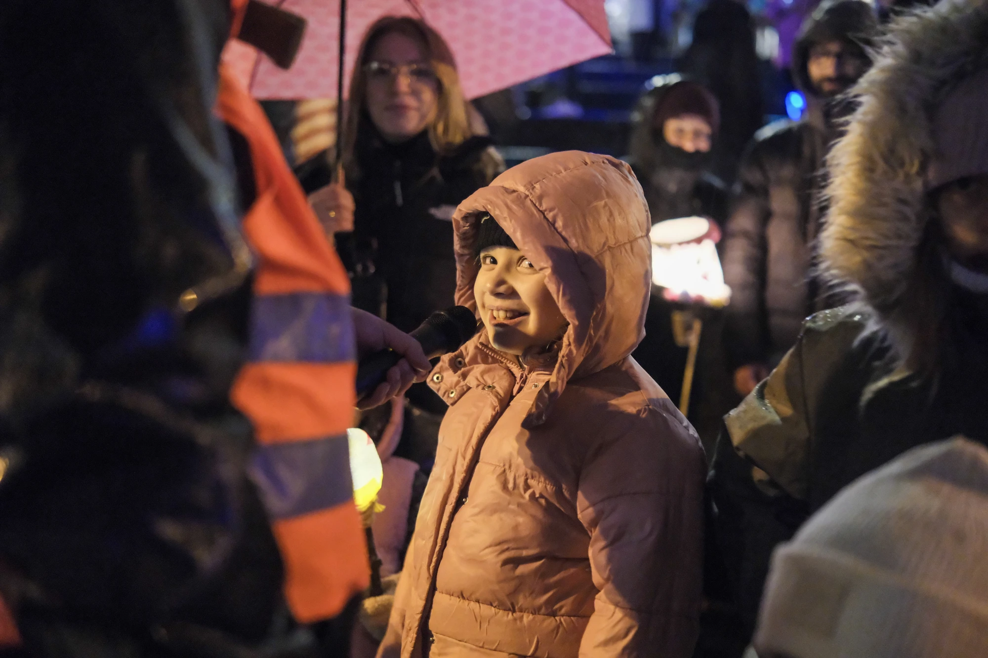 Kind in rosa Winterjacke mit Kapuze steht in einer Menschenmenge bei Nacht, einige halten Laternen und Regenschirme.
