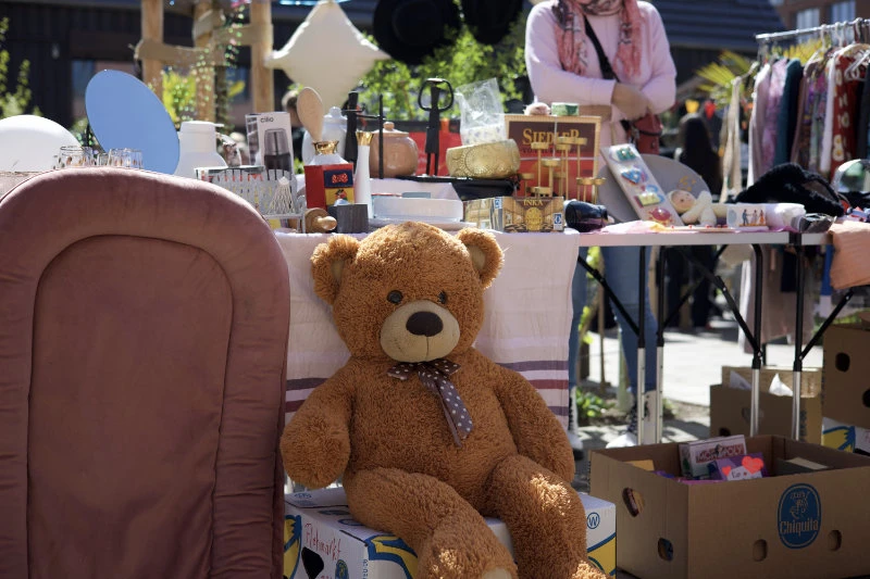 Großer brauner Teddybär mit gepunkteter Schleife sitzt auf einem Karton vor einem Flohmarktstand mit verschiedenen Gegenständen