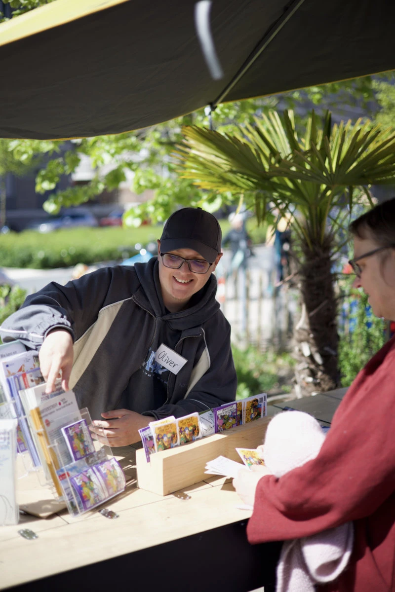 Junger Mann mit Baseballkappe und Namensschild bedient eine Kundin. Er zeigt ihr Programme und Sticker.