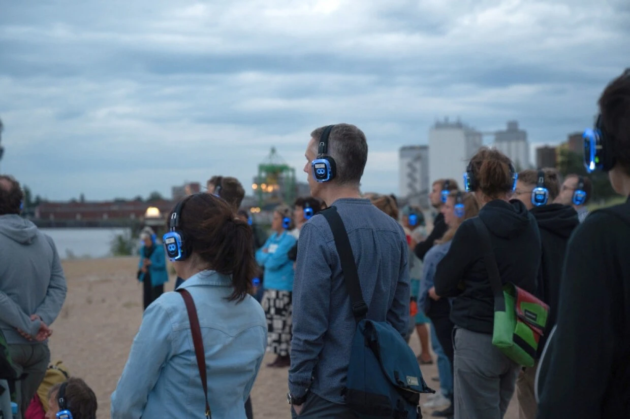 Gruppe von Menschen mit blauen Kopfh&ouml;rern steht am Ufer unter bew&ouml;lktem Himmel