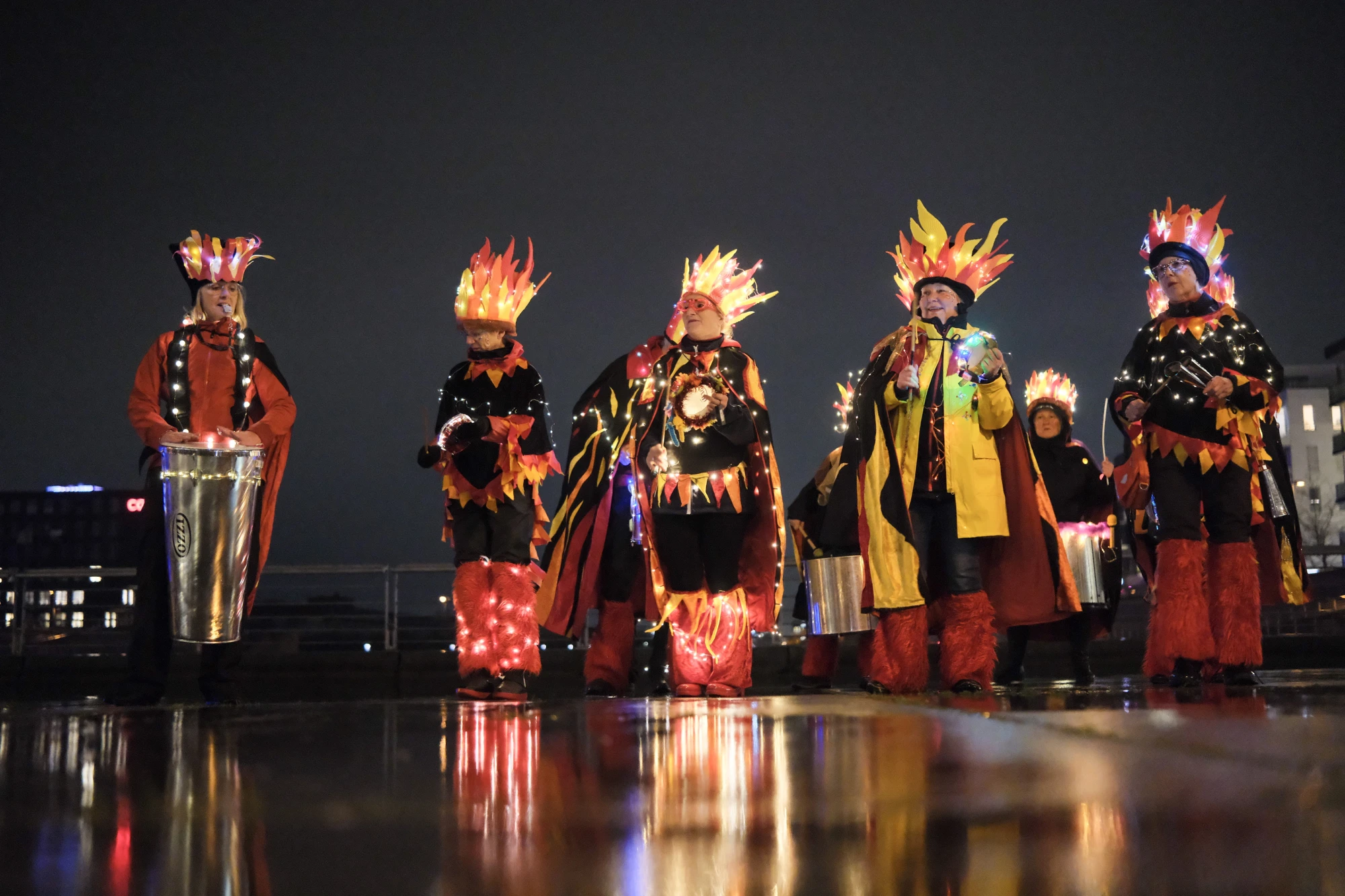 Gruppe von sechs Personen in leuchtenden Kostümen mit Flammenmotiven, eine Person spielt Trommel, nächtliche Stadtkulisse im Hintergrund.