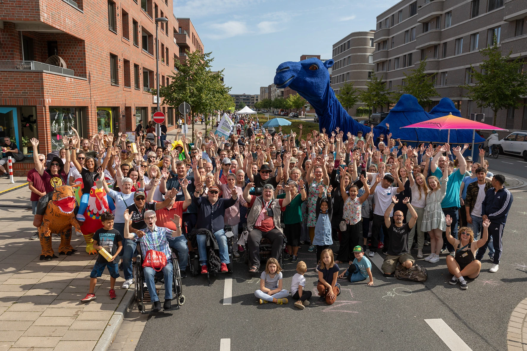 Große Gruppe von Menschen auf einer Straße zwischen modernen Wohngebäuden, viele winken in die Kamera, zwei Personen in Dinosaurierkostümen, mehrere Kinder sitzen vorne auf dem Asphalt