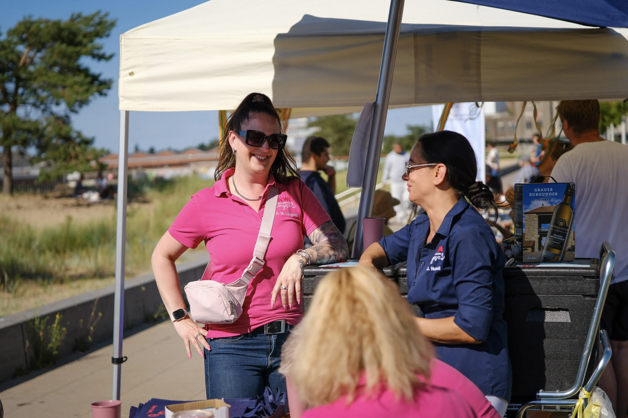 Zwei Frauen unter einem weißen Pavillon im Gespräch, eine trägt ein pinkes Poloshirt und Sonnenbrille, die andere ein dunkelblaues Hemd