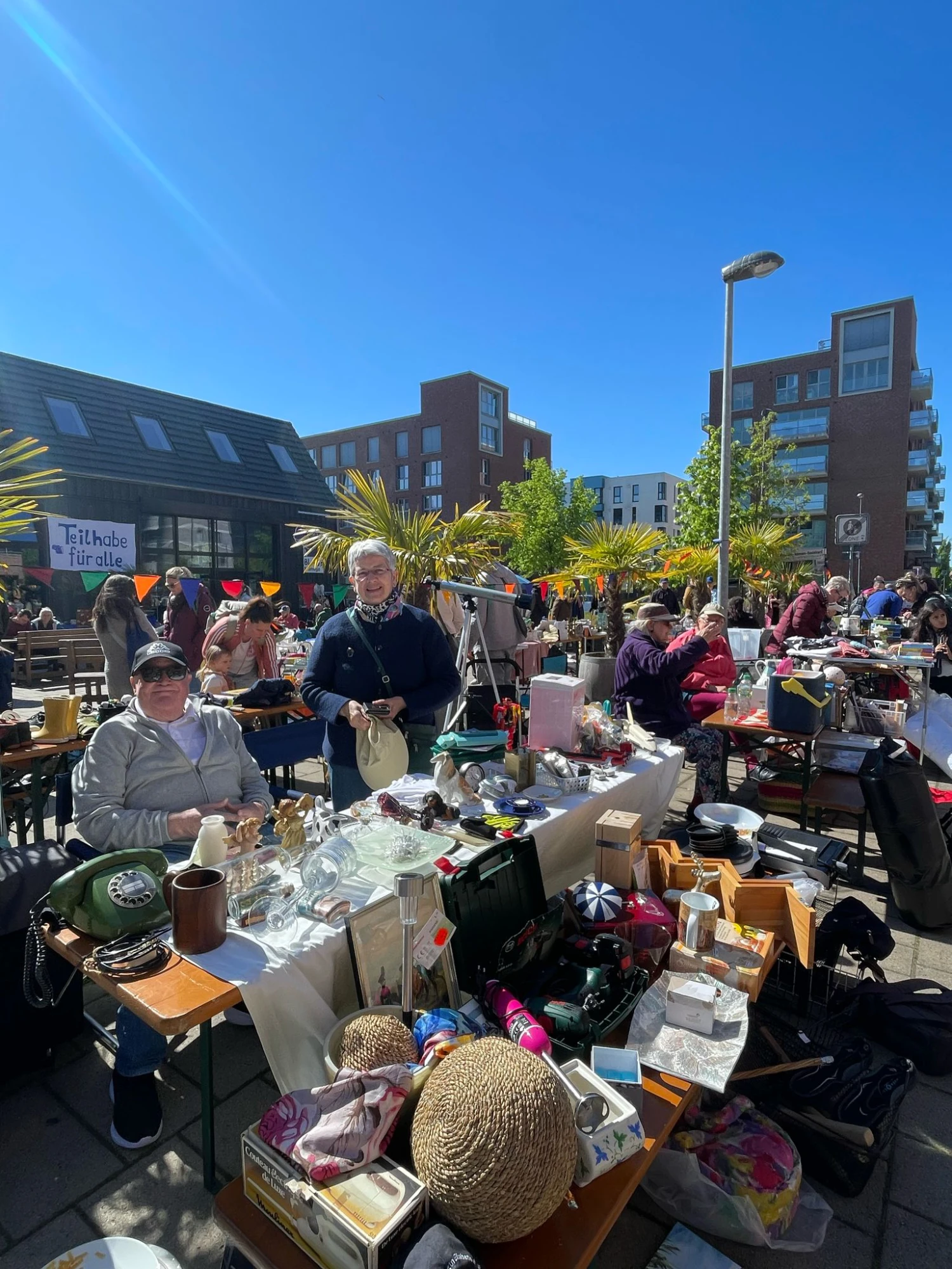 Ein Flohmarkt findet im Freien auf einer gepflasterten Stra&szlig;e statt. Auf mehreren Tischen liegen verschiedene Gegenst&auml;nde wie Geschirr, Gl&auml;ser, B&uuml;cher, Taschen, K&ouml;rbe und Dekorationsartikel. Menschen stehen und sitzen an den Tischen, einige betrachten die Waren. Im Hintergrund sind moderne Wohngeb&auml;ude, B&auml;ume und ein Schild mit der Aufschrift 'T&uuml;tchen f&uuml;r alle' zu sehen. Die Szene ist von Sonnenlicht und blauem Himmel gepr&auml;gt.