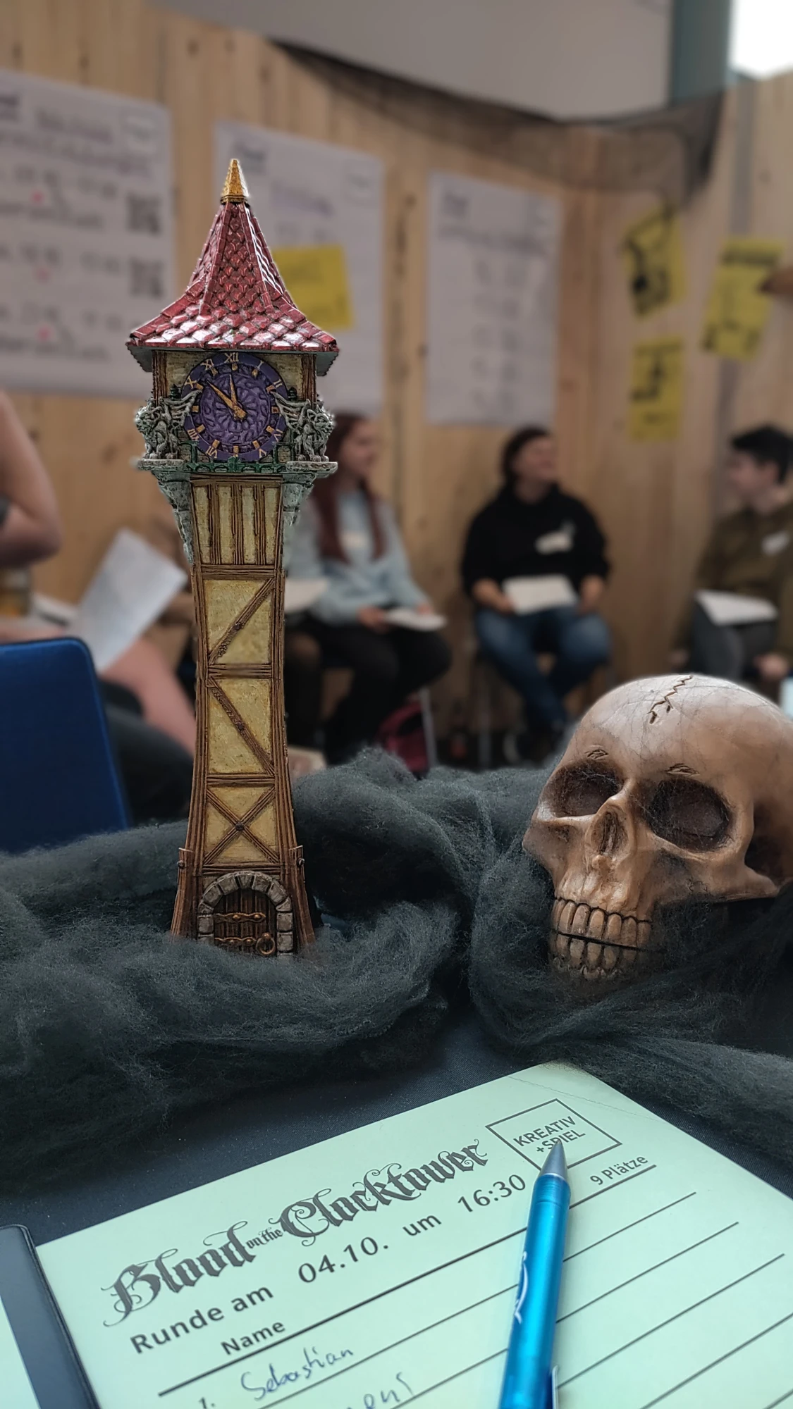 Eine Turmfigur steht auf einem Tisch, daneben ein Totenkopf. Der Turm ist das Symbol von dem Spiel Blood on the Clocktower. Vor dem Turm und dem Totenkopf ist ein Zettel und Stift, wo Mitspieler notiert sind. Im Hintergrund sind verschwommen einige Personen zu sehen, die im Halbkreis sitzen.