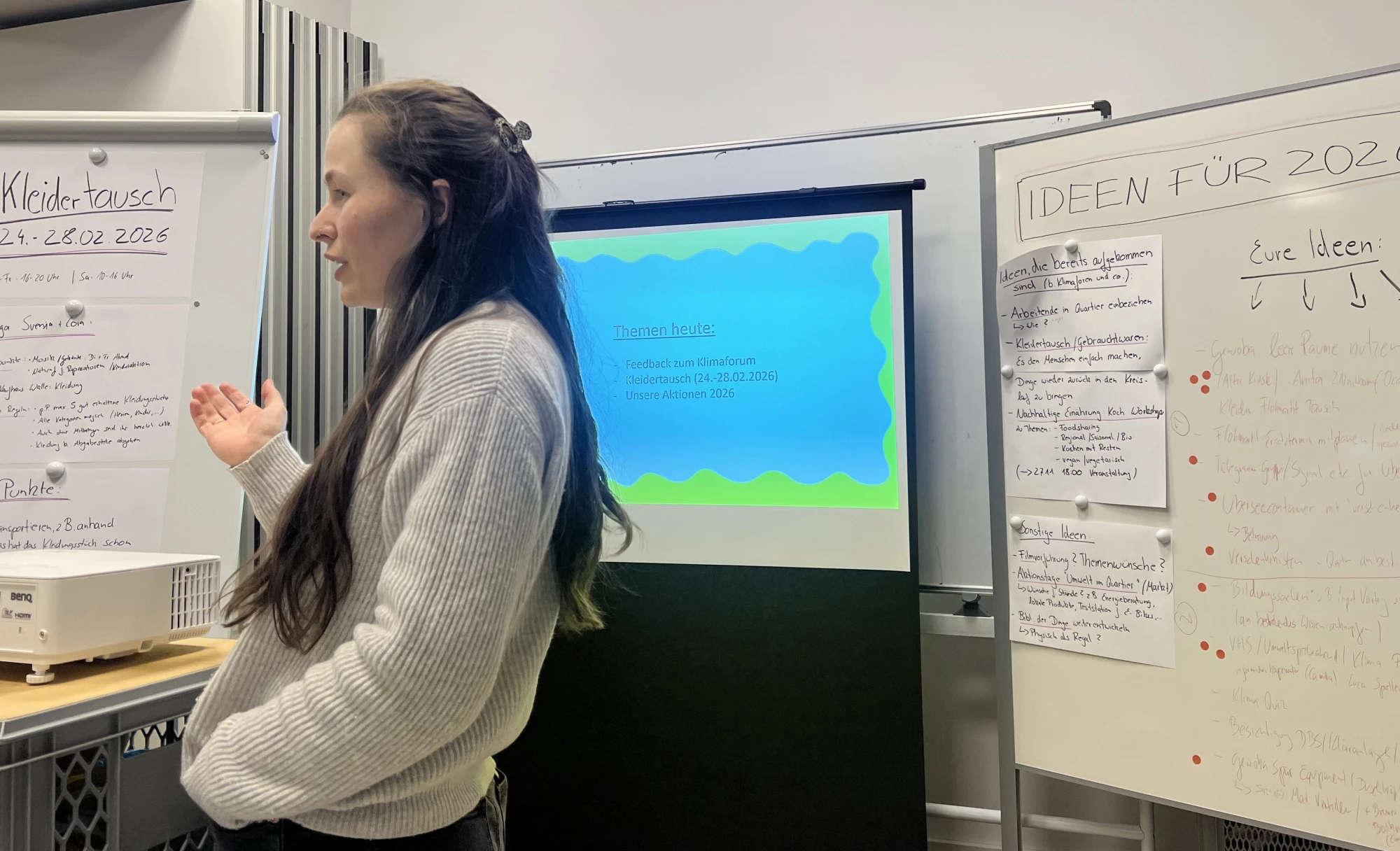 Frau mit langem Haar und weißem Pullover steht vor Flipcharts und einem Projektorbildschirm in einem Besprechungsraum