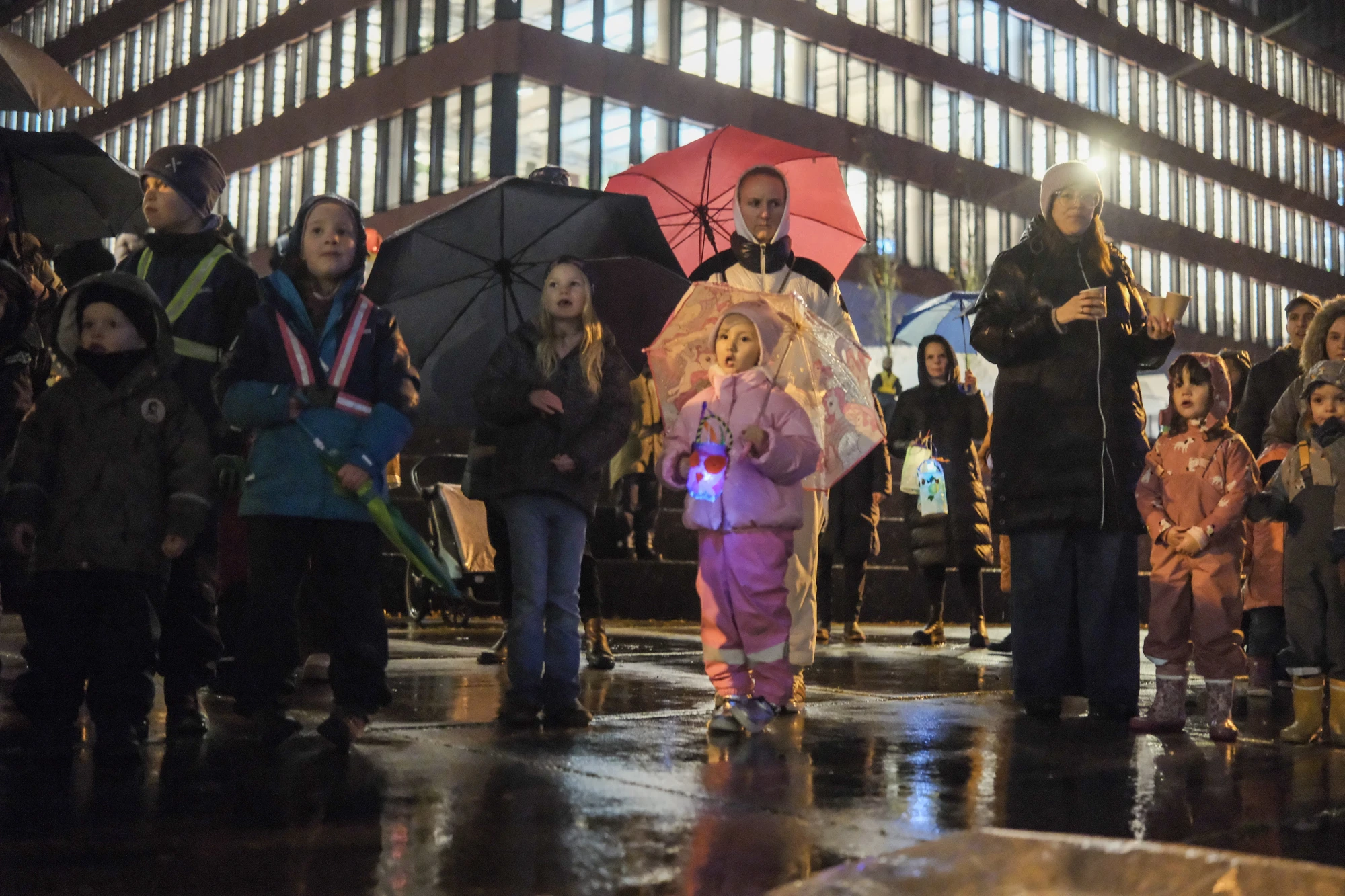Gruppe von Kindern und Erwachsenen mit Regenschirmen und Laternen bei Nacht auf nassem Boden vor beleuchtetem Gebäude.