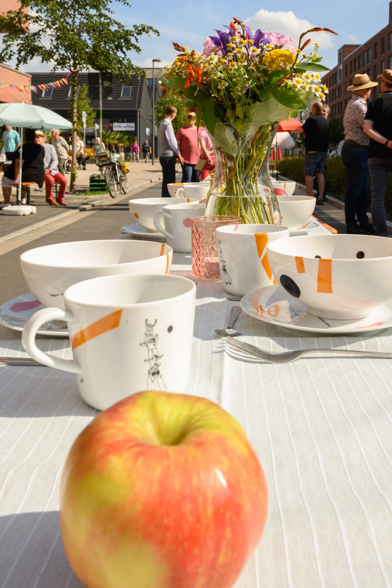 Tisch mit weißem Geschirr mit orangefarbenen Mustern, Apfel im Vordergrund und Blumenstrauß in Vase bei sonnigem Straßenfest mit Menschen im Hintergrund