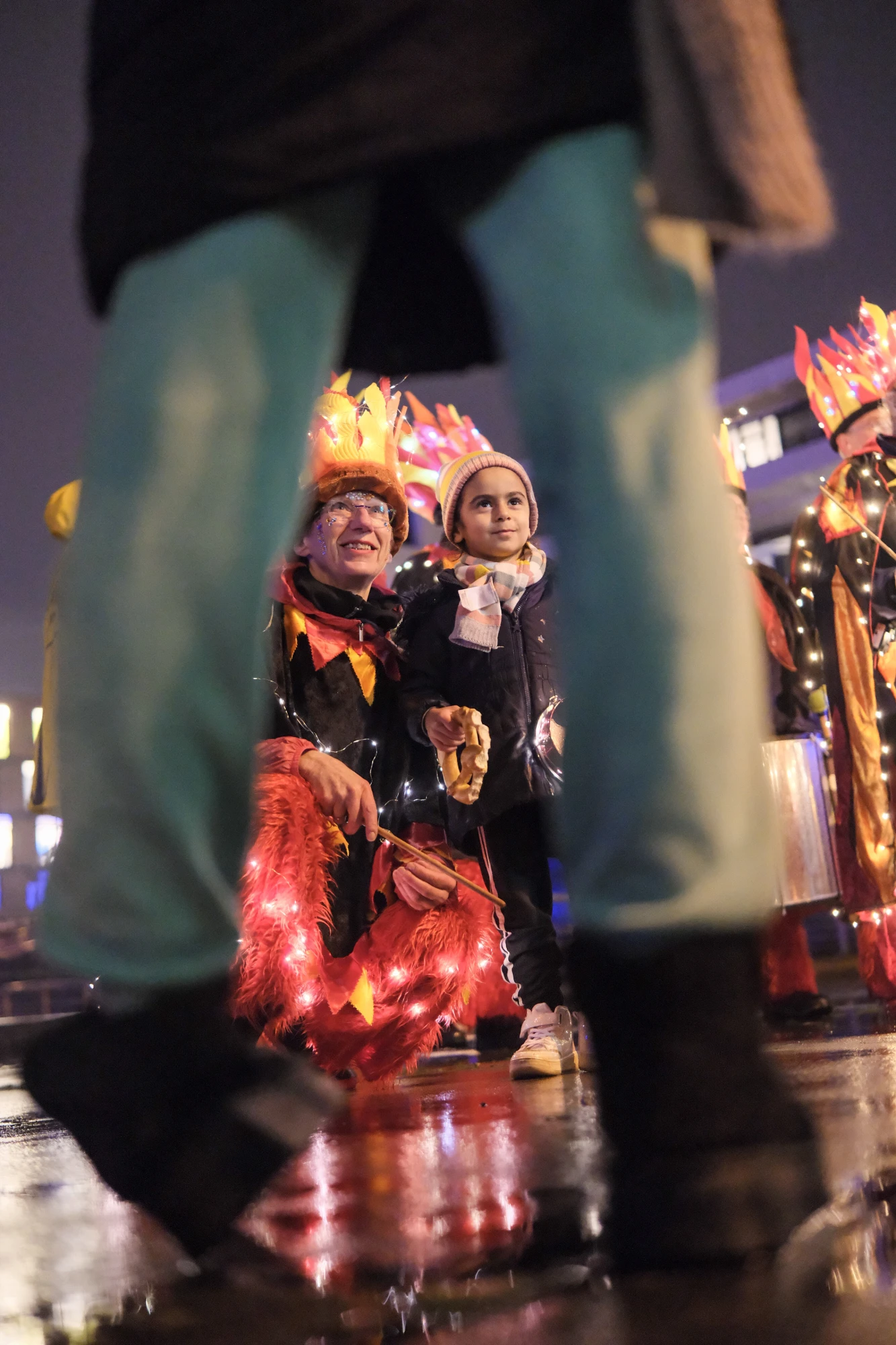 Mehrere Personen mit leuchtenden, bunten Laternen in Form von Flammen bei Nacht, im Vordergrund Beine und Füße einer weiteren Person.