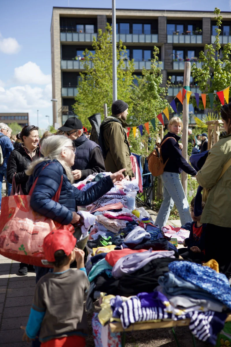 Bunte Wimpelgirlanden hängen über mehreren Tischen, die mit Kleidung und Textilien bedeckt sind. Menschen stehen dicht beieinander, betrachten die ausgelegten Sachen und greifen nach einzelnen Kleidungsstücken. Im Hintergrund sind moderne Gebäude mit großen Fenstern und Balkonen sowie grüne Bäume zu sehen. Die Szene spielt im Freien bei Tageslicht