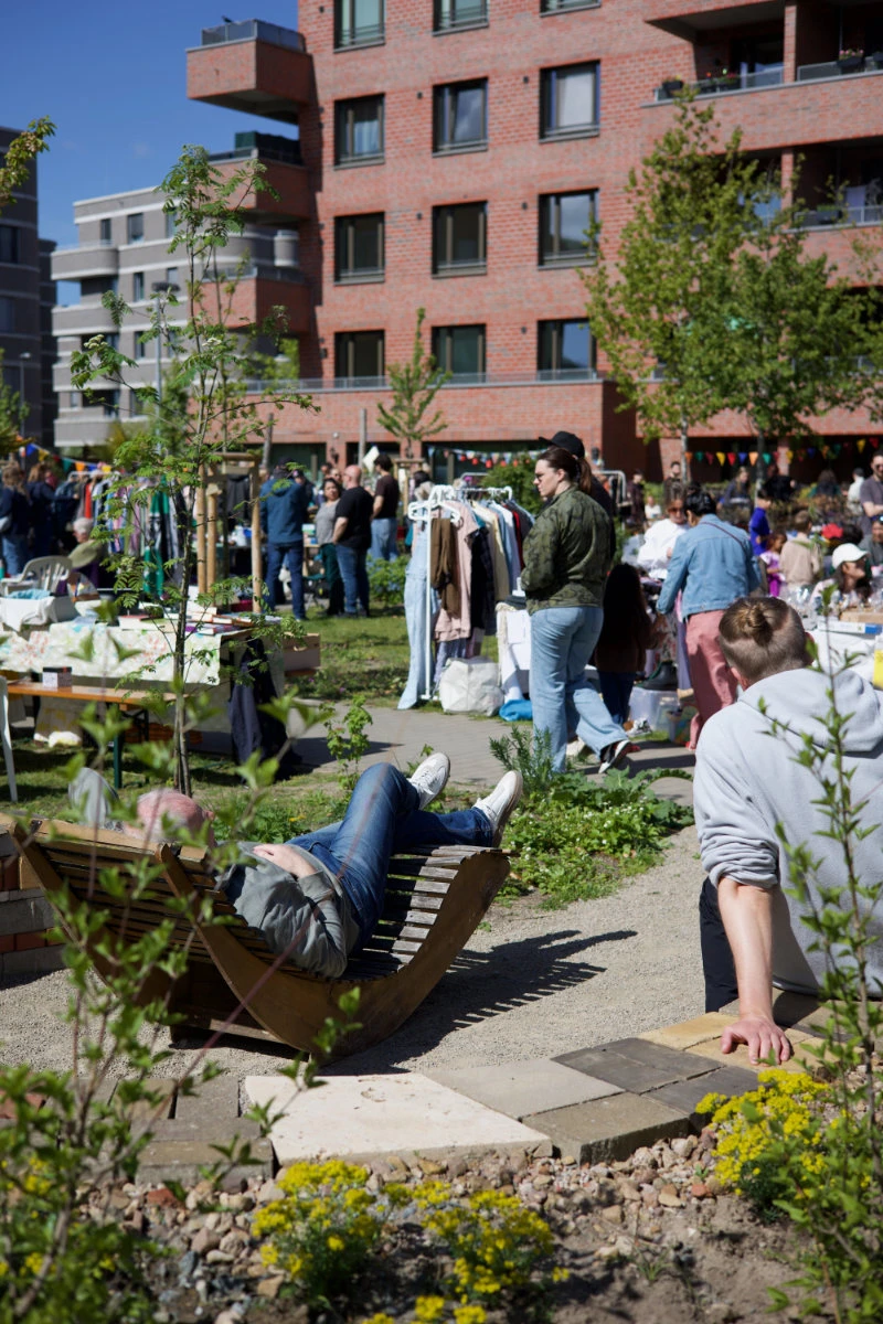 11.	Im Vordergrund entspannt eine Person auf einer geschwungenen Holzliege, während eine weitere Person am Rand eines Kieswegs sitzt. Im Hintergrund findet ein Flohmarkt im Freien statt. Mehrere Menschen stehen oder gehen zwischen Tischen und Kleiderständern, auf denen verschiedene Gegenstände und Kleidung angeboten werden. Die Szene spielt in einem begrünten Innenhof, umgeben von modernen Wohngebäuden aus rotem Backstein. Bäume und Sträucher spenden Schatten, und die Atmosphäre wirkt lebendig und sommerlich.