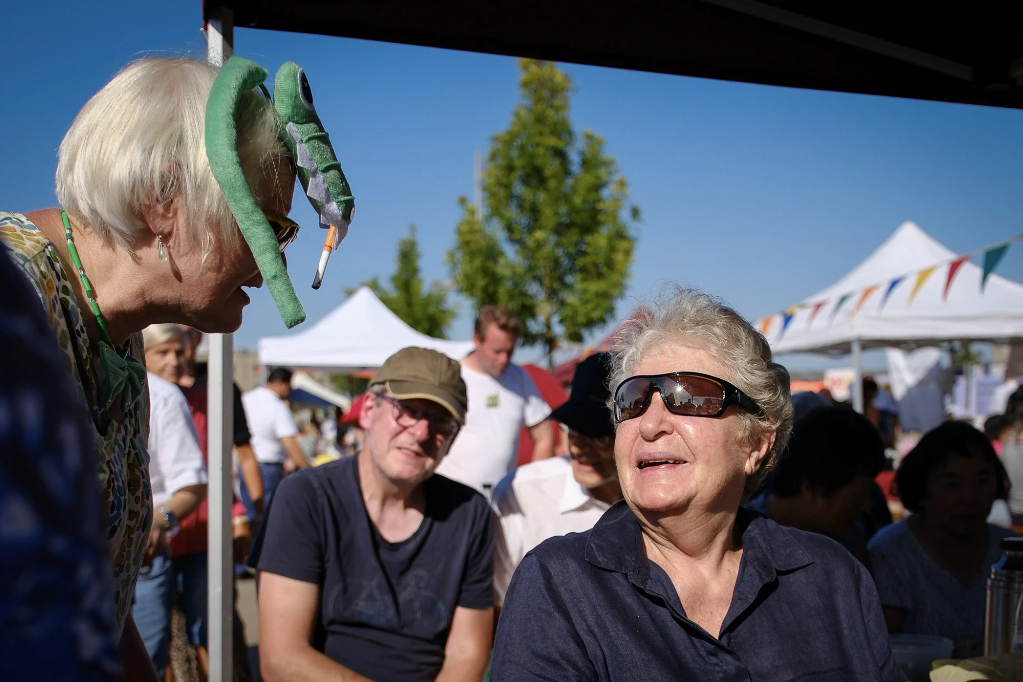Eine ältere Frau mit Sonnenbrille schaut eine andere Frau an. Im Hintergrund sitzen weitere Menschen und es sind Pavillons zu sehen.