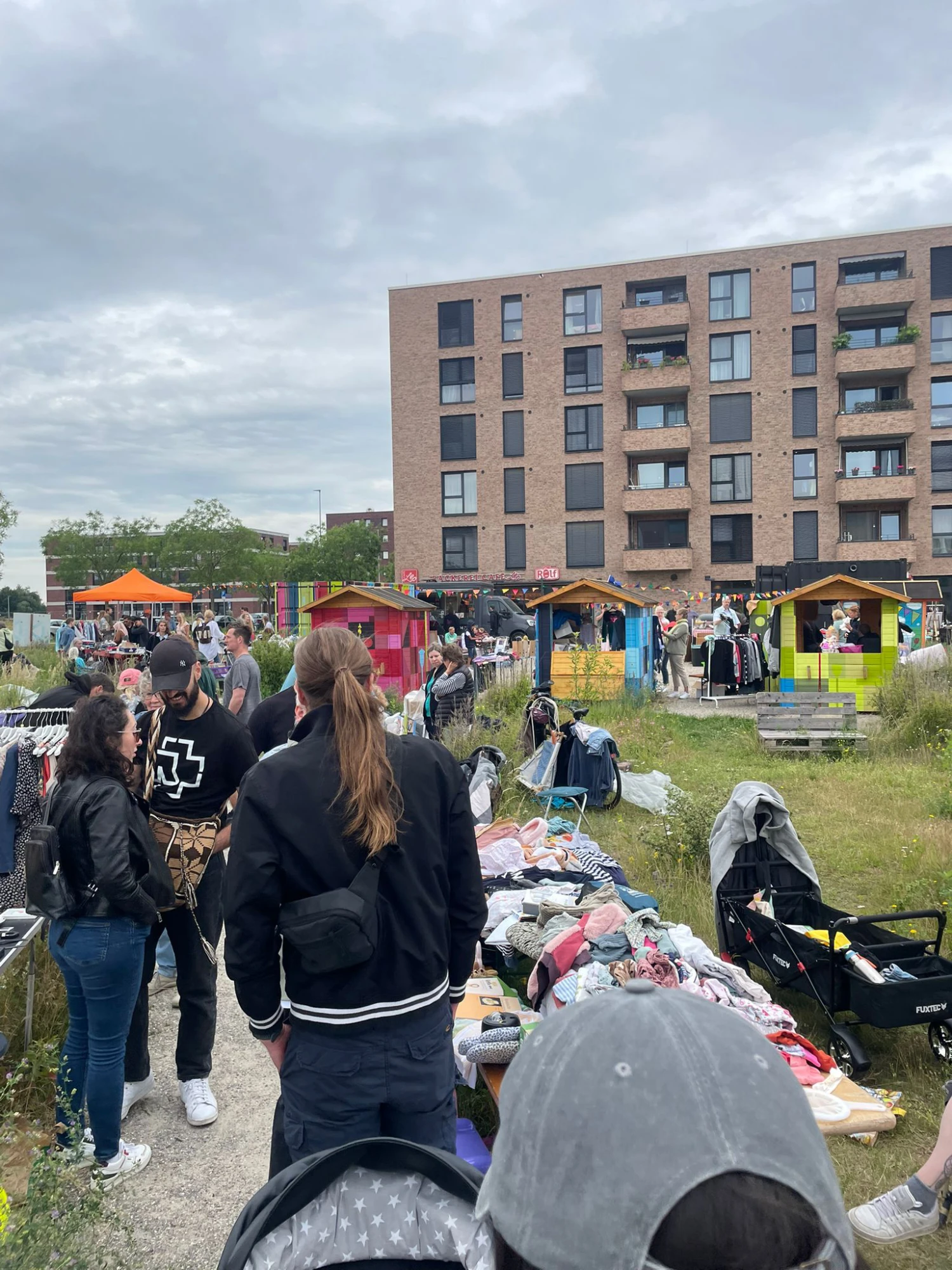 Menschen auf einem Flohmarkt mit Kleidung und anderen Waren auf Tischen und in K&ouml;rben vor einem mehrst&ouml;ckigen Wohngeb&auml;ude bei bew&ouml;lktem Himmel