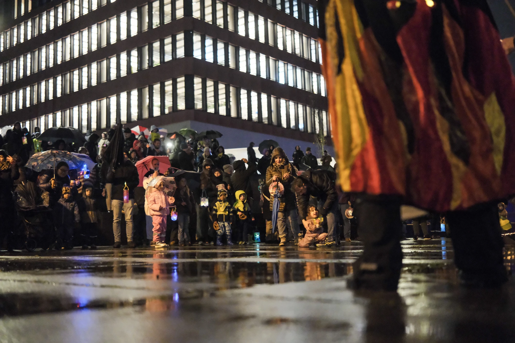 Menschen mit Regenschirmen und Laternen stehen bei Nacht auf nassem Asphalt vor einem Gebäude mit beleuchteten Fenstern.