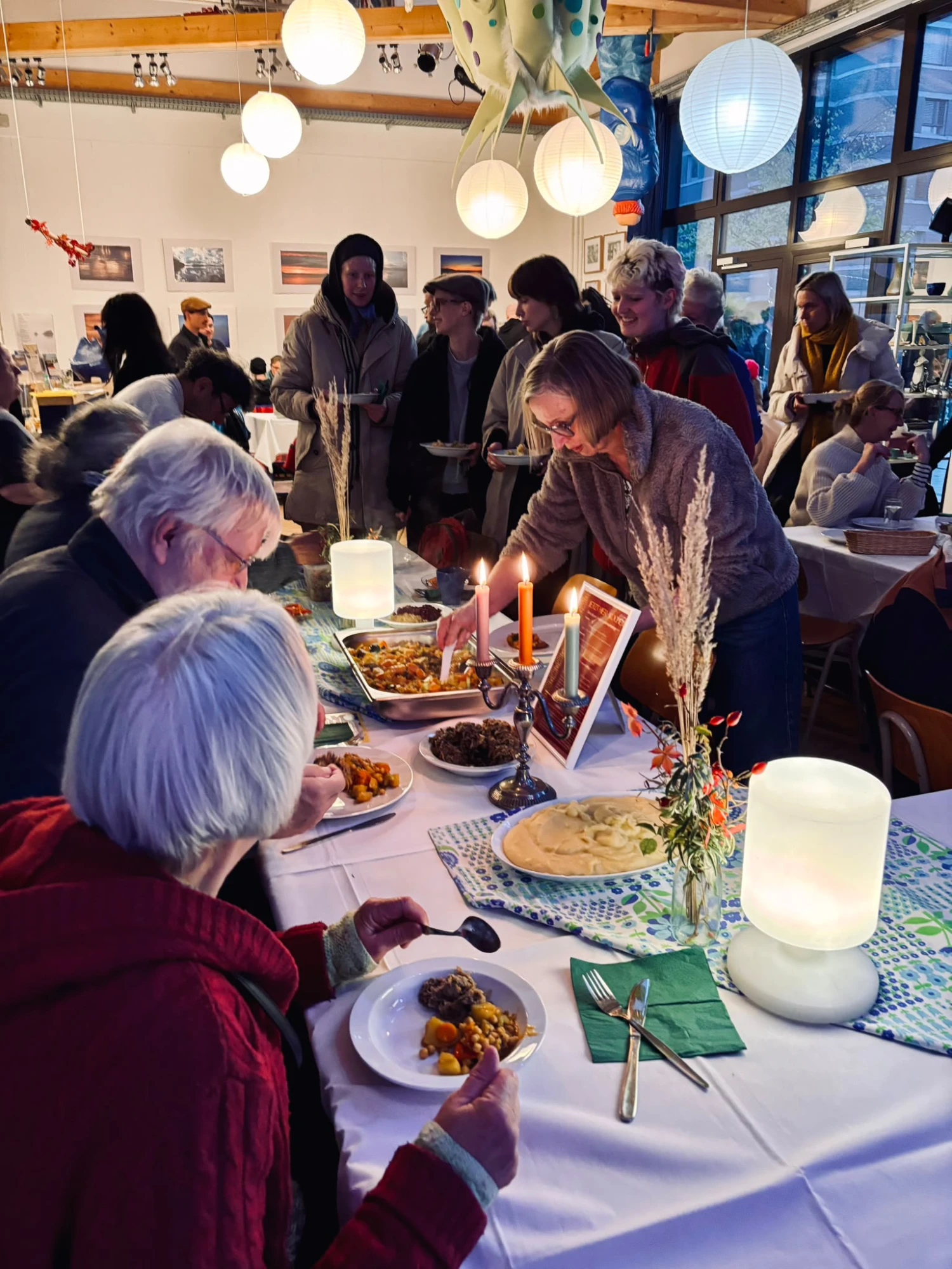 Menschen sitzen am gedeckten Tisch, andere Menschen sehen und nehmen sich Essen auf den Teller.