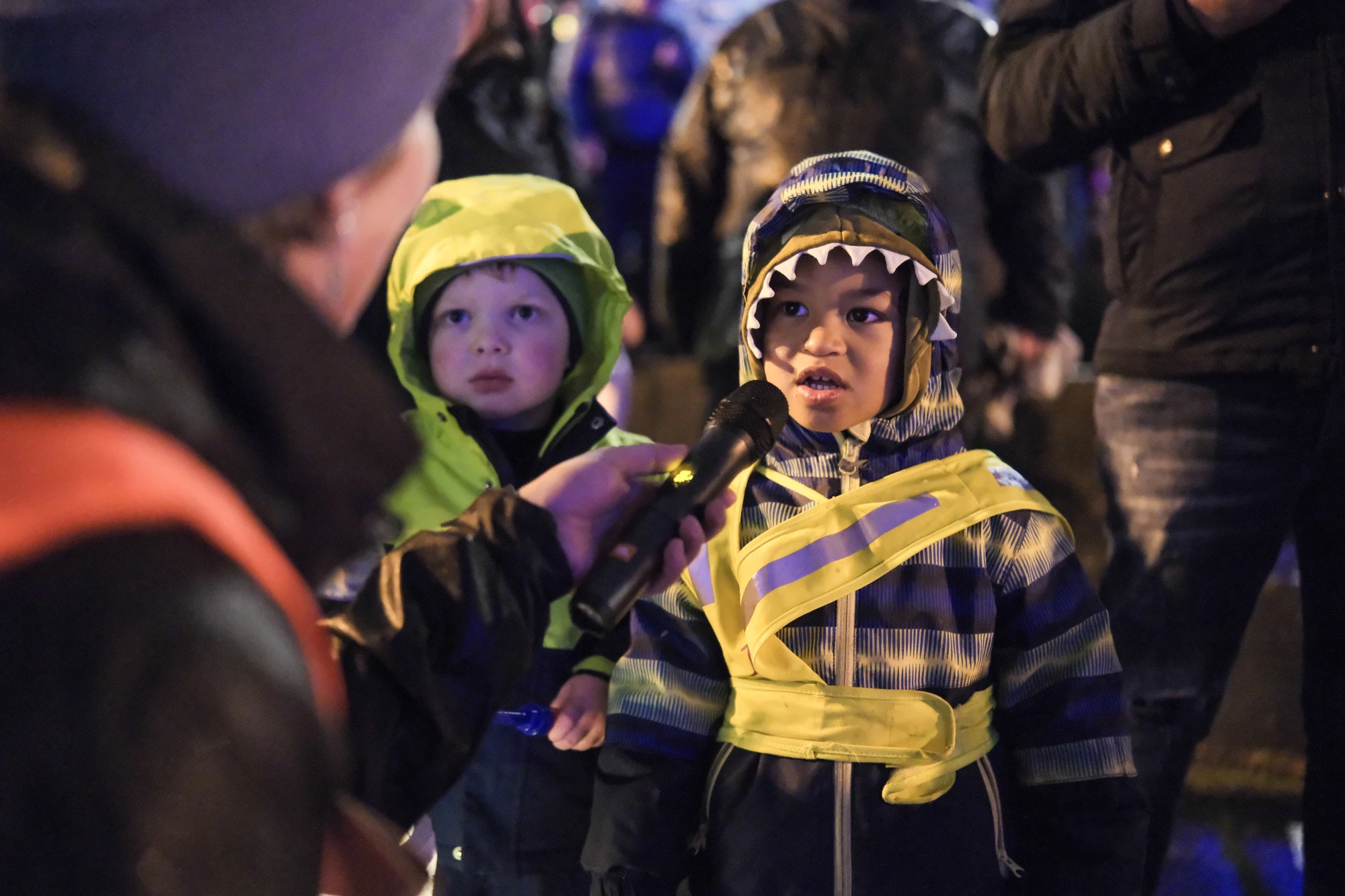 Kind in rosa Jacke und Gummistiefeln steht auf nassem Boden, umgeben von mehreren Personen, eine hält einen Regenschirm.
