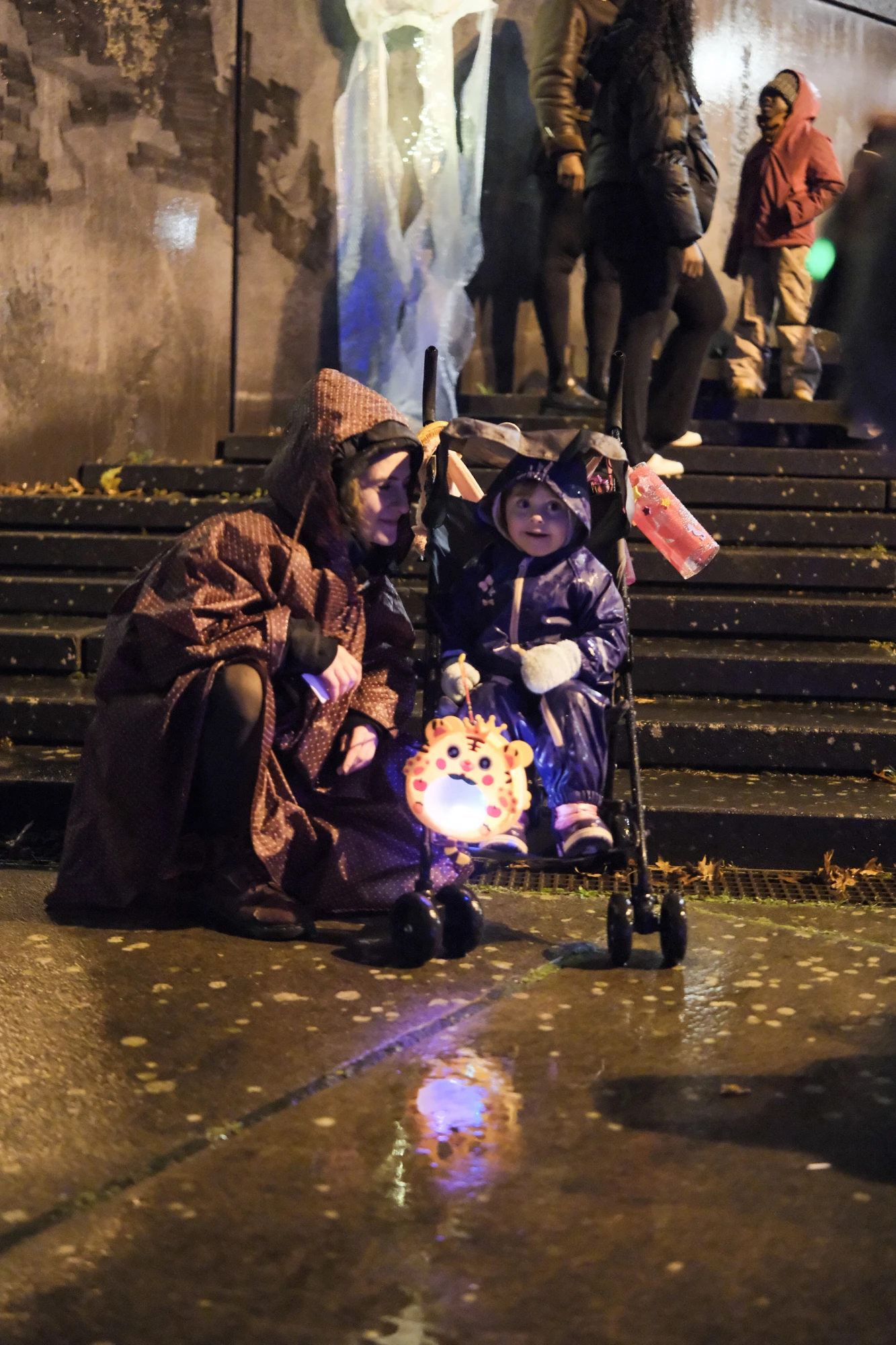 Zwei Kinder in Regenmänteln sitzen auf einer Treppe, eines hält eine leuchtende Laterne mit Gesichtsmotiv, im Hintergrund stehen mehrere Personen.