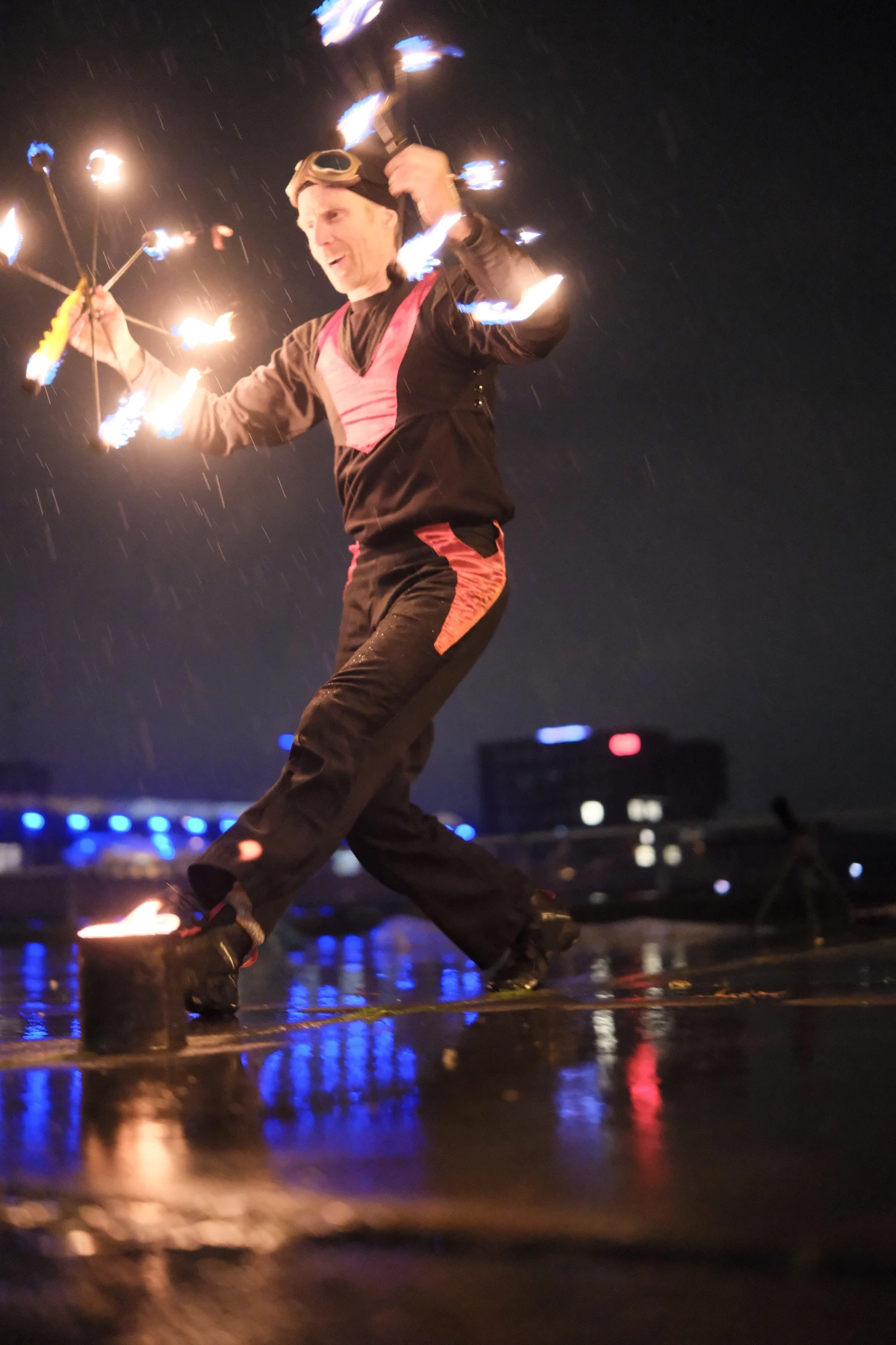 Person in schwarzem Outfit mit roten Akzenten jongliert nachts mit zwei brennenden Feuerreifen auf nassem Boden vor dunklem Hintergrund.