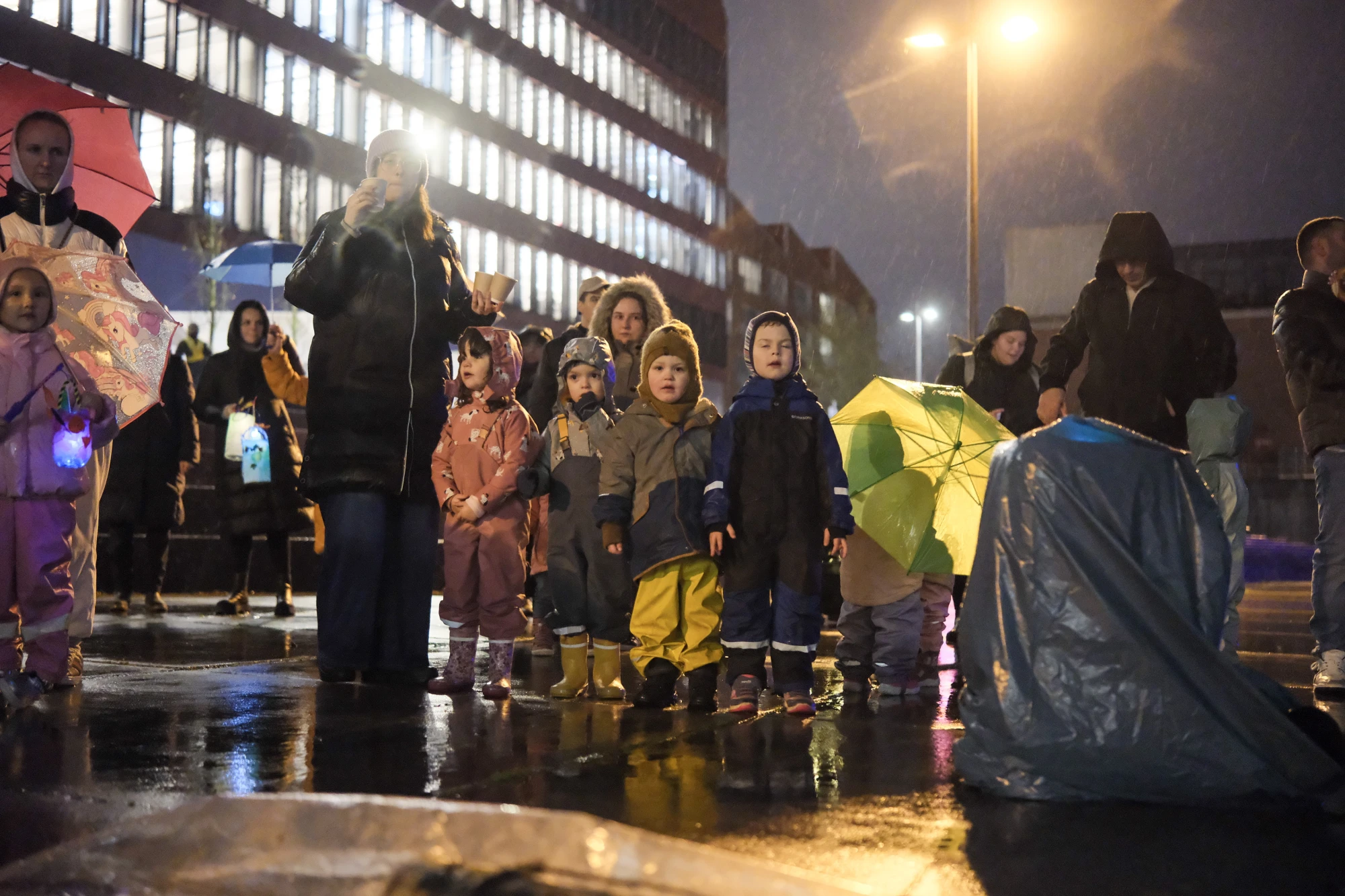 Mehrere Personen, darunter Kinder mit reflektierenden Warnwesten, stehen auf nassem Boden bei Nacht vor einem beleuchteten Gebäude.