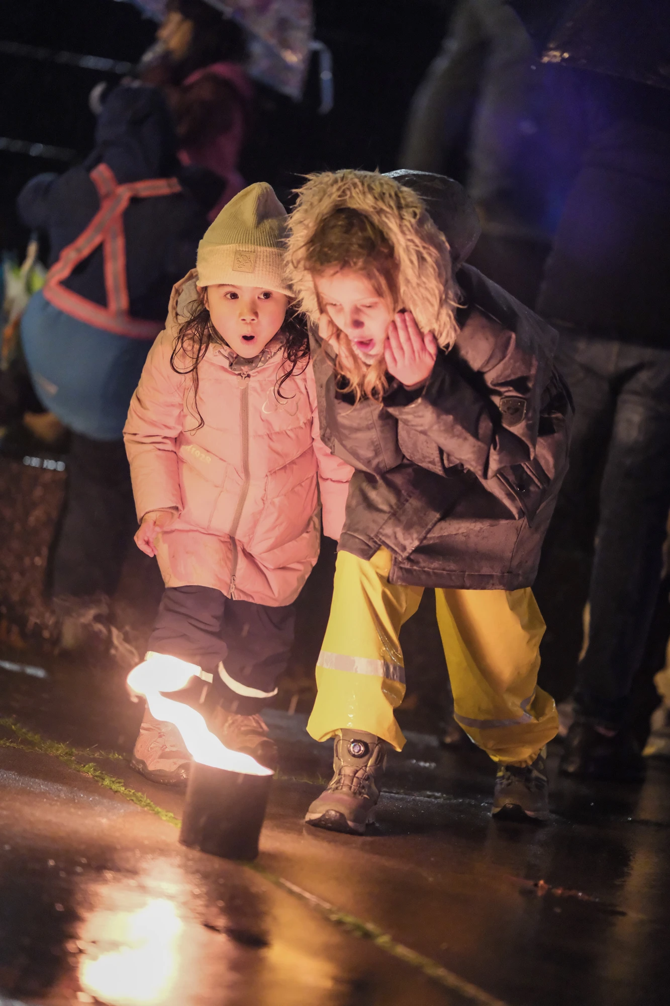 Zwei Kinder in Winterjacken und Mützen bücken sich über eine kleine brennende Laterne auf nassem Boden.