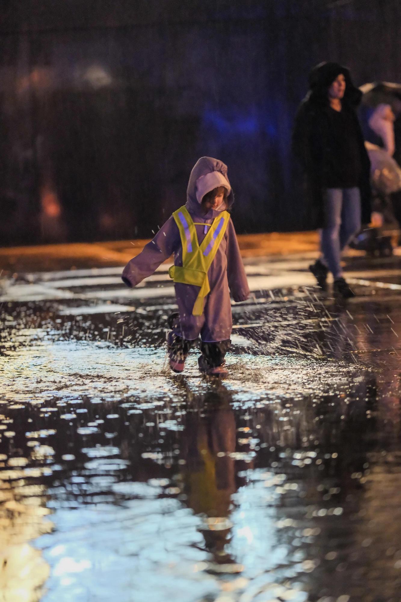 Kind mit Kapuzenjacke und gelber Warnweste läuft auf nasser Straße mit Reflexionen bei Nacht.