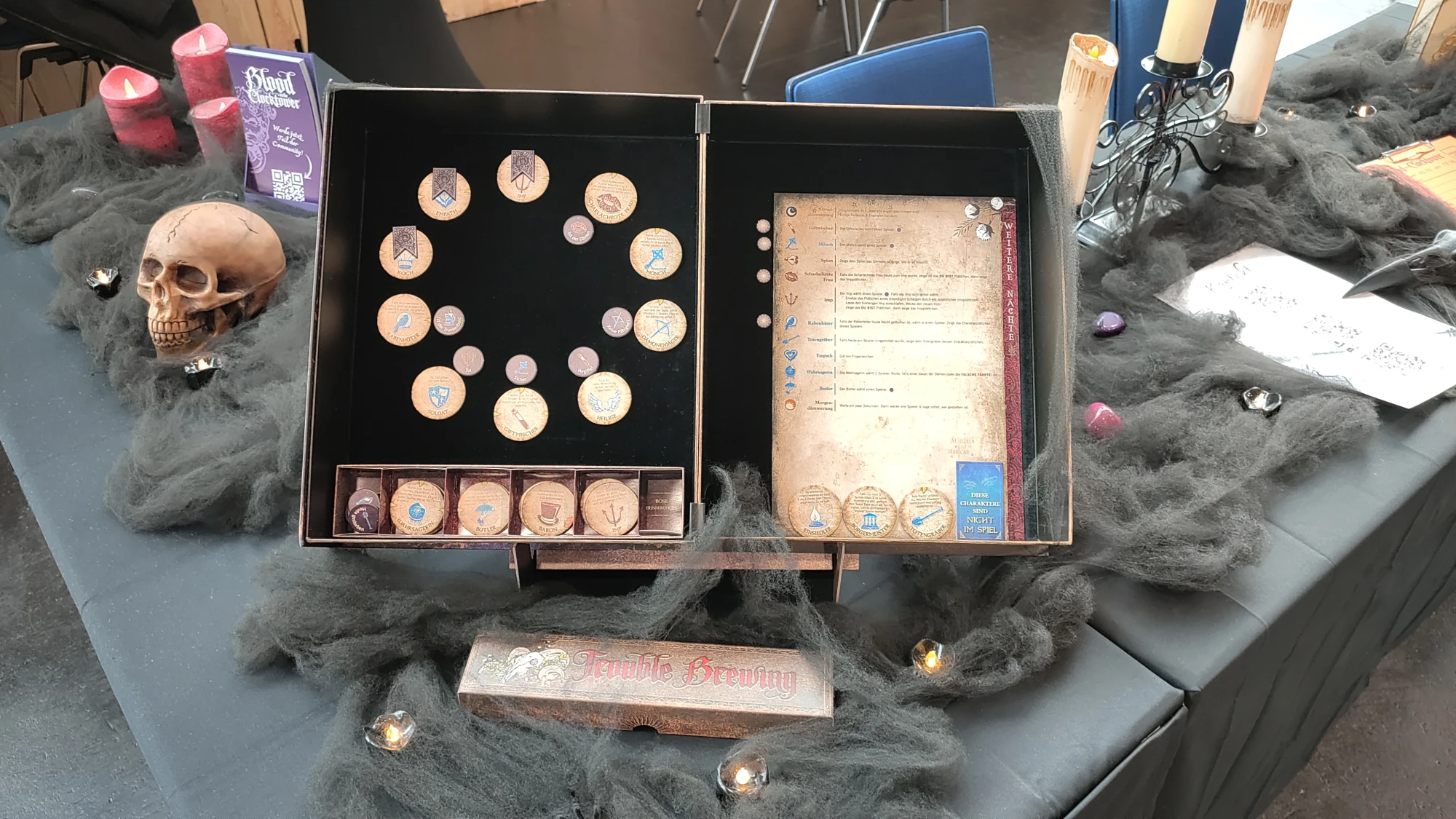 Ein dekorativer Spieletisch mit einem geöffneten Spielkasten des Gesellschaftsspiels Blood on the Clocktower. Der Kasten zeigt verschiedene Holzmarker mit Symbolen und Beschriftungen, sowie eine Übersichtstafel mit Spielregeln. Um den Kasten herum liegen schwarze Deko-Spinnweben, kleine LED-Teelichter, ein künstlicher Schädel, rote Kerzen und ein Kerzenständer mit weißen Kerzen, die eine mystische, Halloween-ähnliche Atmosphäre erzeugen.