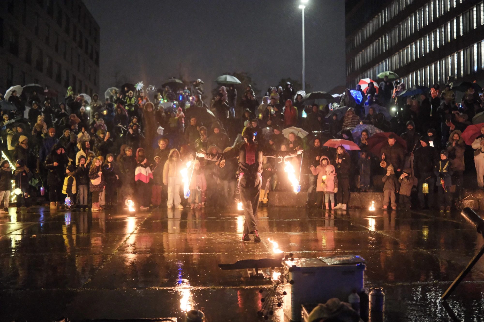 Feuerkünstler in Bewegung vor einer großen Menschenmenge bei Nacht, die mit Regenschirmen und warmen Kleidern zuschaut.