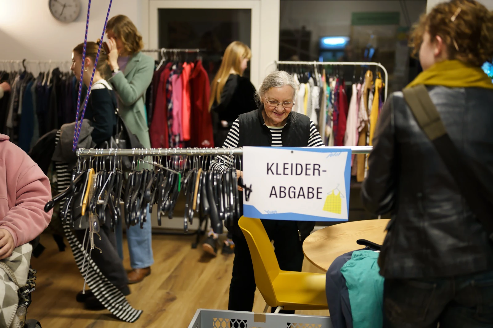 Personen stehen in einem Raum mit Kleiderständern und einem Schild mit der Aufschrift 'KLEIDER-ABGABE'