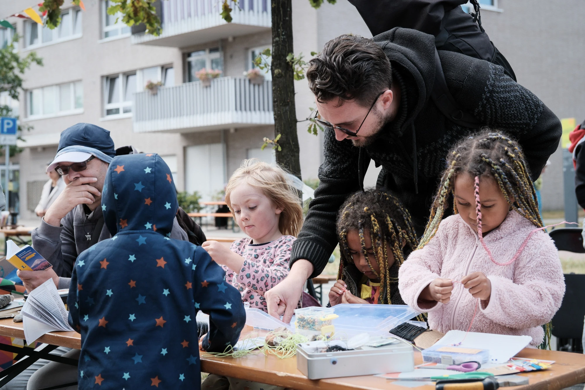 Ein Erwachsener mit dunkler Jacke beugt sich über einen Tisch im Freien und zeigt mehreren Kindern etwas auf dem Tisch. Die Kinder tragen bunte Kleidung, darunter ein Kapuzenpullover mit Sternenmuster und geflochtene Zöpfe. Auf dem Tisch liegen Bastelmaterialien wie Papier, Stifte und Schnüre. Im Hintergrund sind ein Baum, ein mehrstöckiges Wohnhaus mit Balkonen und weitere Personen zu sehen.