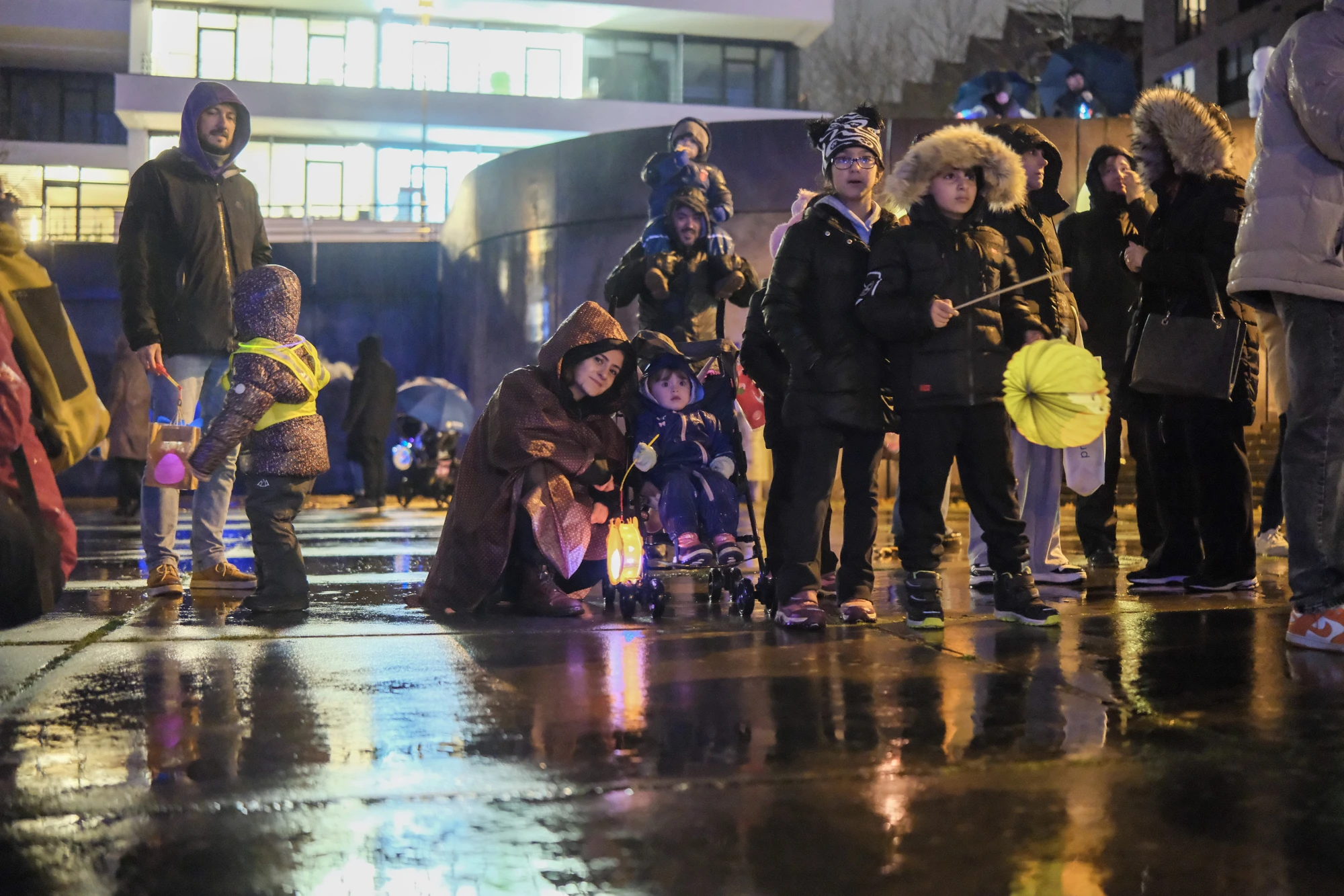 Gruppe von Kindern und Erwachsenen mit Regenschirmen und Laternen bei Nacht auf nassem Boden vor beleuchtetem Gebäude.