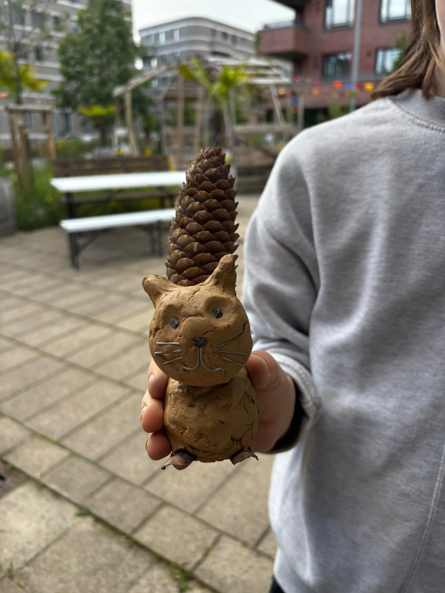 Hand hält eine kleine Figur aus Ton mit einem Zapfen als Schwanz, die wie ein Tier mit Gesicht und Schnurrhaaren aussieht