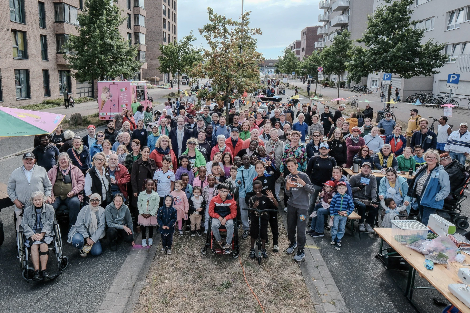 Eine große Gruppe von Menschen steht auf einer gesperrten Straße zwischen mehrstöckigen Wohnhäusern. Viele Personen lächeln und blicken in Richtung Kamera. Einige sitzen im Rollstuhl, andere stehen oder hocken. Im Vordergrund sind Kinder, Erwachsene und ältere Menschen zu sehen. Rechts steht ein Tisch mit technischen Geräten und Papieren. Im Hintergrund sind Bäume, geparkte Autos und weitere Menschen erkennbar. Die Szene wirkt wie ein Nachbarschaftstreffen oder Straßenfest.