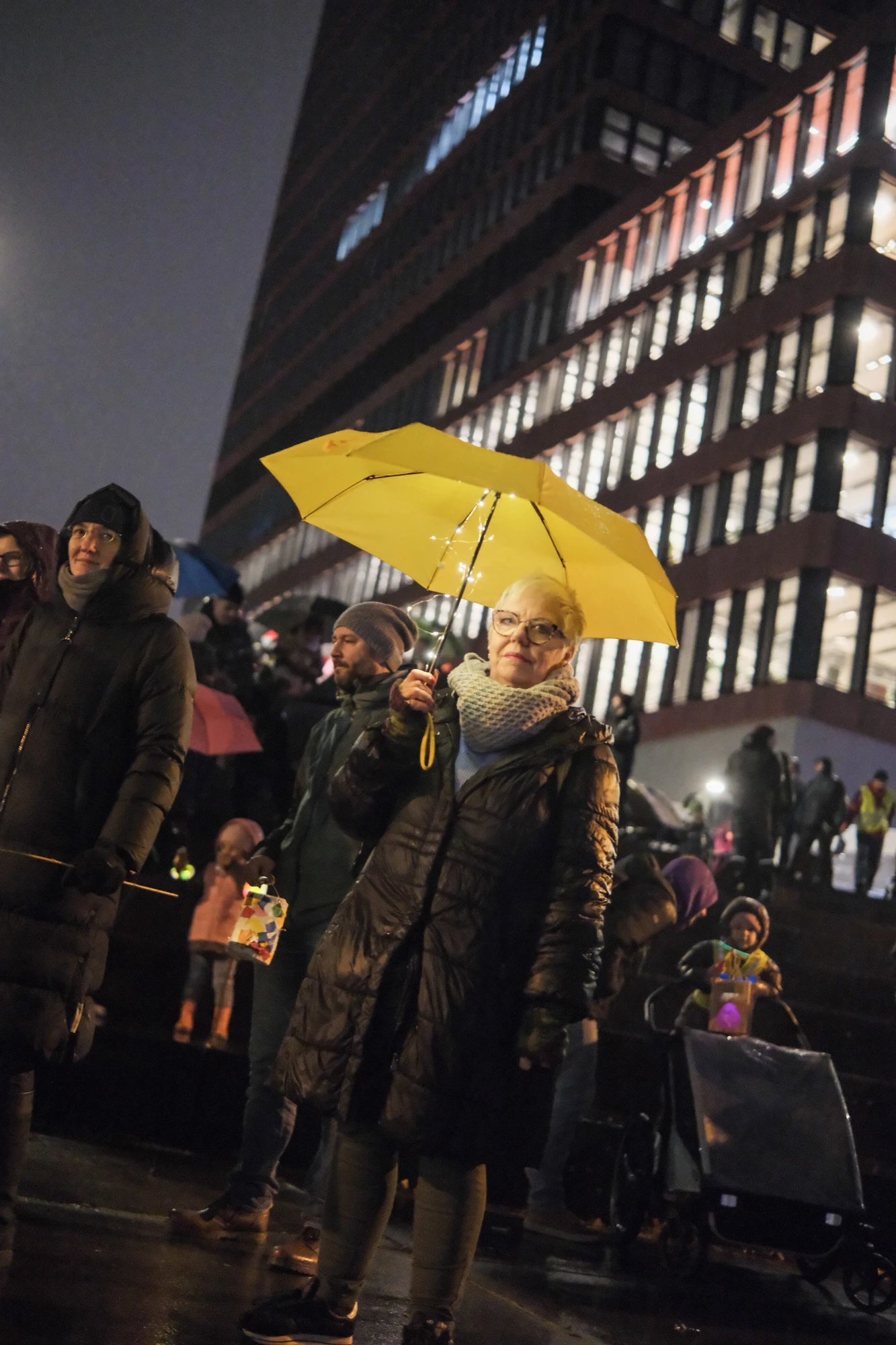 Person mit gelbem Regenschirm und dunklem Mantel steht vor einem beleuchteten Hochhaus in der Nacht, umgeben von mehreren Menschen.