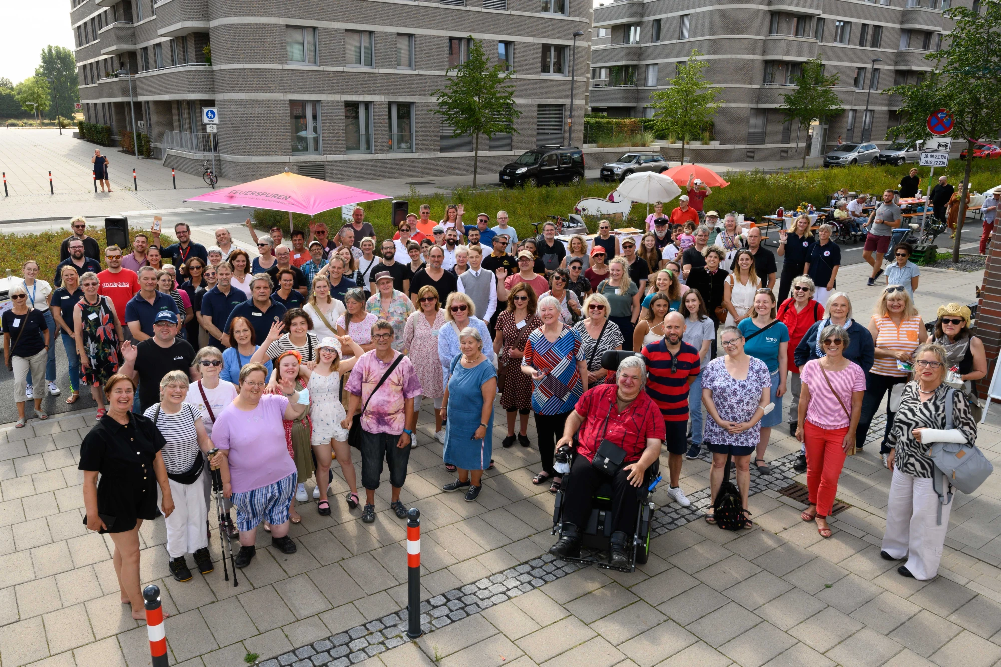 Große Gruppe von Menschen unterschiedlichen Alters steht auf einem gepflasterten Platz vor modernen Wohngebäuden, einige halten bunte Regenschirme