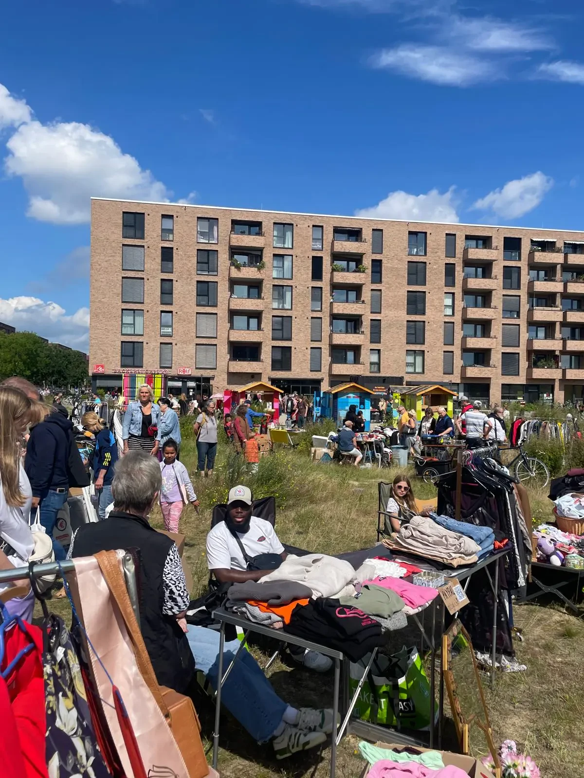 Menschen auf einem Flohmarkt mit Tischen voller Kleidung und einem mehrst&ouml;ckigen Wohngeb&auml;ude im Hintergrund