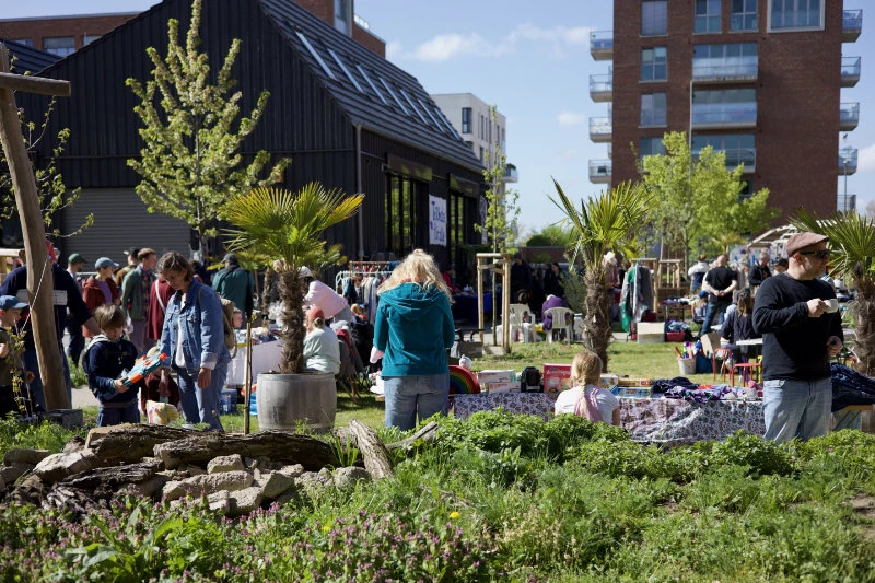 Menschen verschiedener Altersgruppen stehen und sitzen bei sonnigem Wetter in einem urbanen Garten mit Pflanzen und einem schwarzen Holzgebäude im Hintergrund