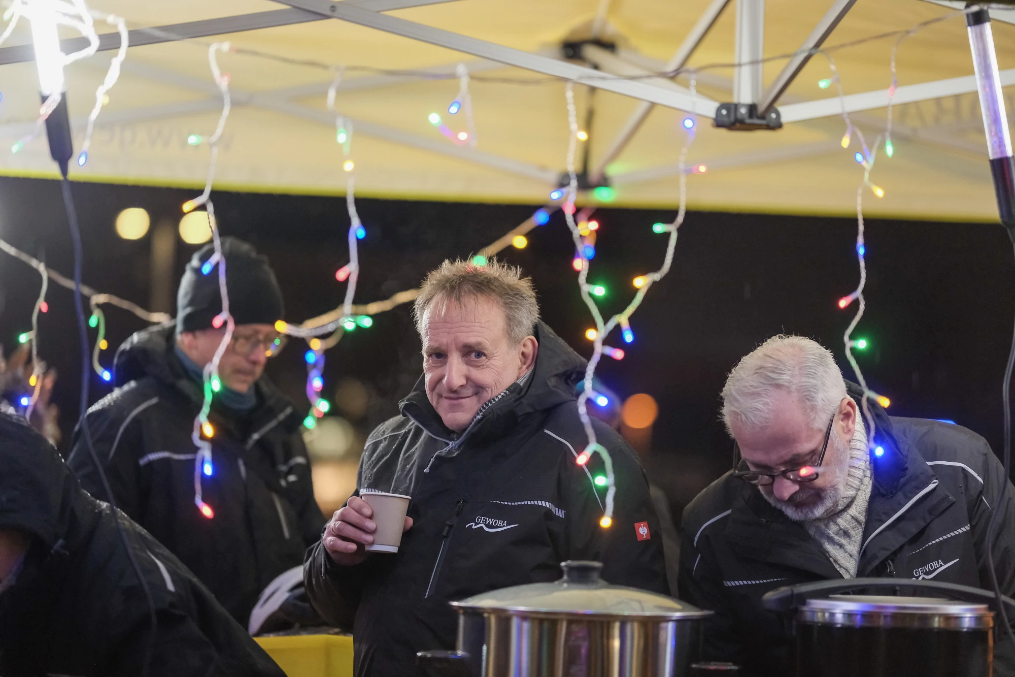 Mehrere Personen in dunklen Jacken stehen unter einem gelben Zelt mit bunten Lichterketten, eine Person hält einen Becher in der Hand.