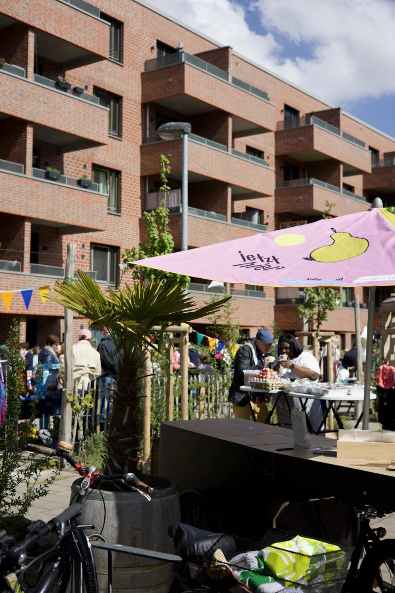 Mehrstöckiges Wohngebäude mit Balkonen und Menschen, die an einem sonnigen Tag an einem Marktstand mit rosa Sonnenschirm und Dekorationen stehen