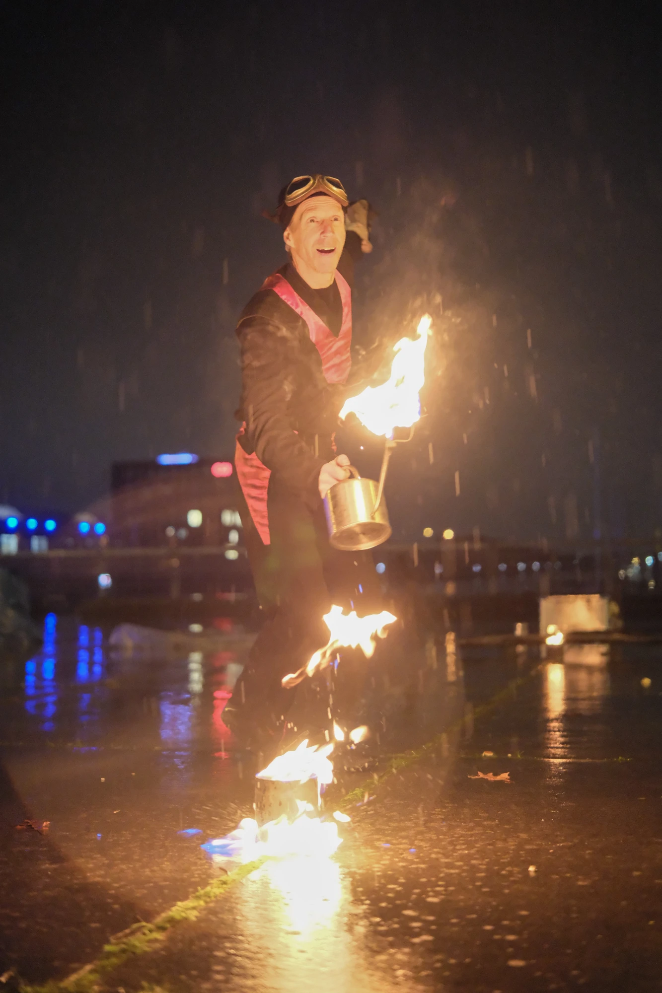 Person in dunkler Kleidung mit reflektierenden Streifen hält brennende Laterne über nassem Boden bei Nacht.
