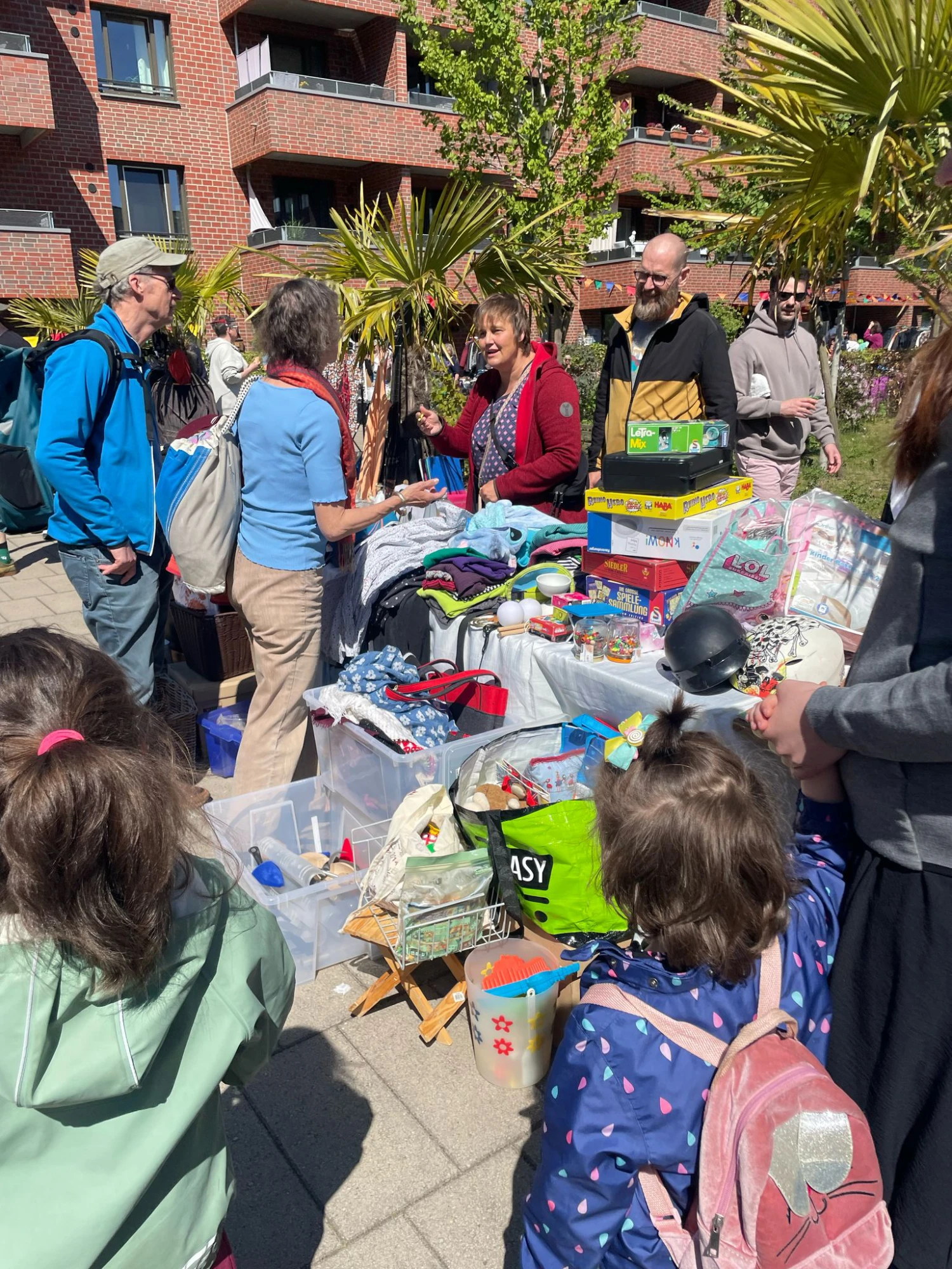Mehrere Personen stehen an einem Tisch mit verschiedenen Kleidungsst&uuml;cken, Spielzeug und Haushaltsgegenst&auml;nden im Freien vor einem Wohngeb&auml;ude.