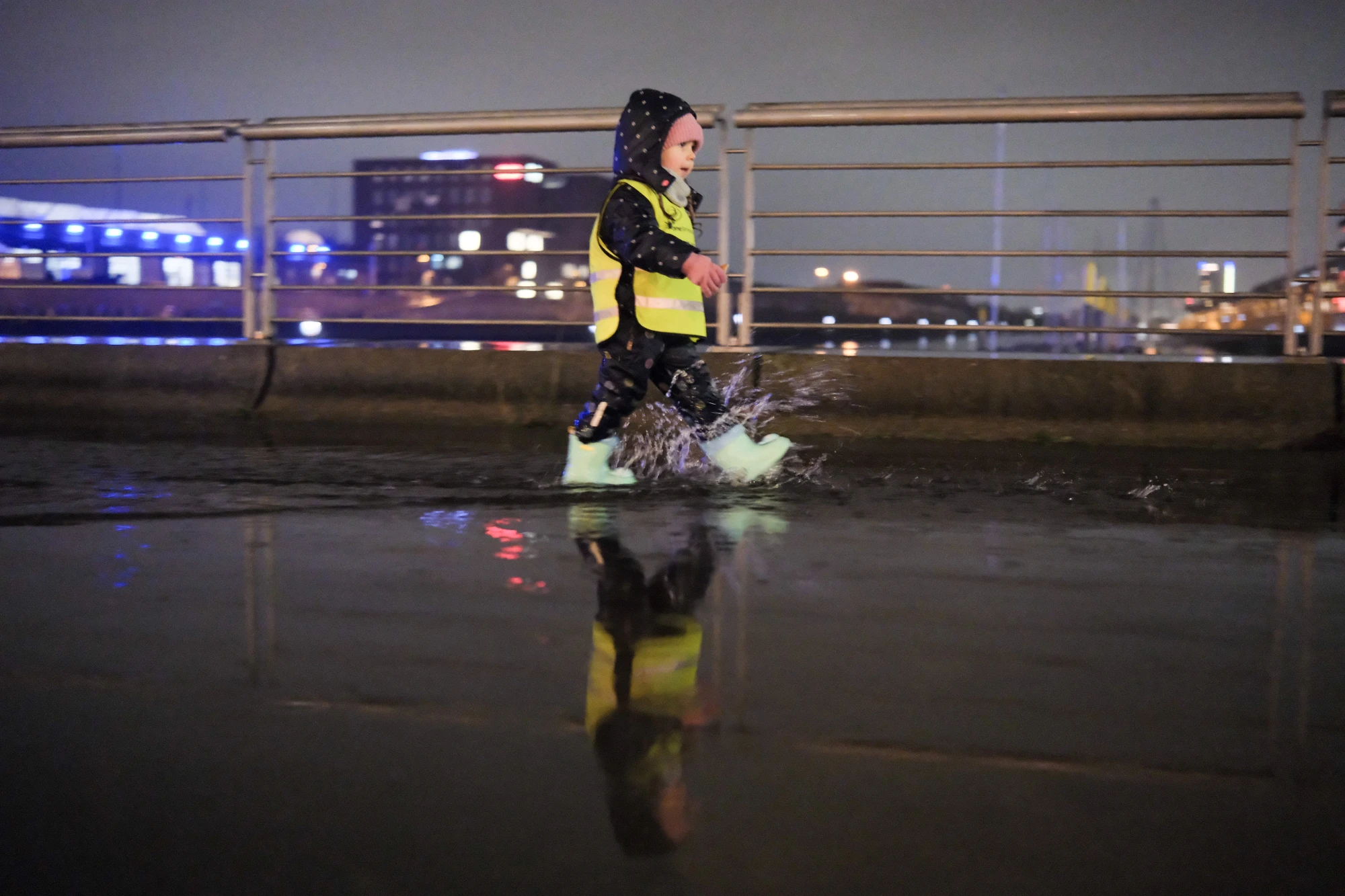 Kind mit Kapuzenjacke und gelber Warnweste läuft auf nasser Straße mit Reflexionen bei Nacht.