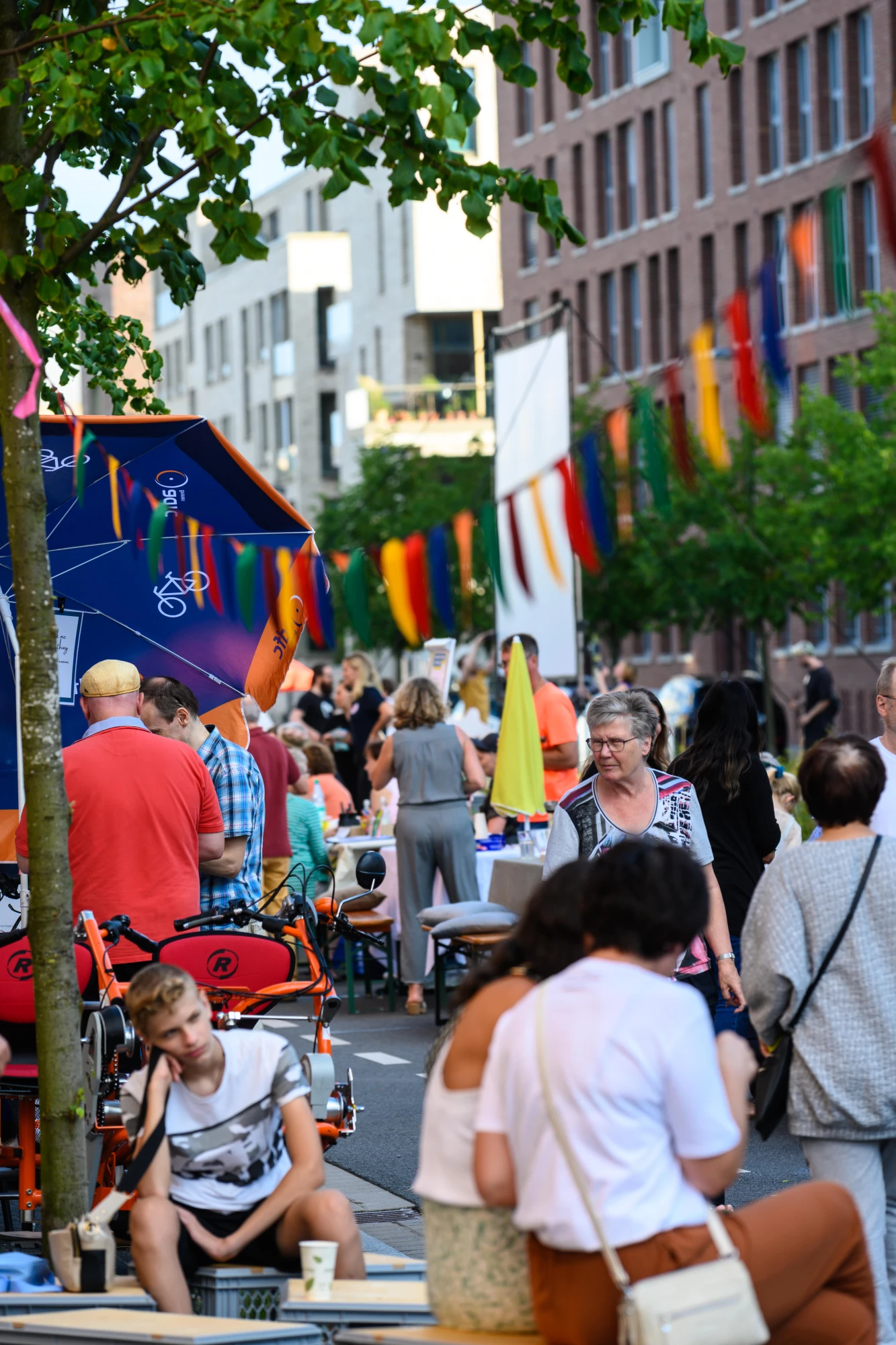 Straßenszene mit Menschen, die an Tischen sitzen und stehen, bunte Wimpelketten hängen zwischen Gebäuden