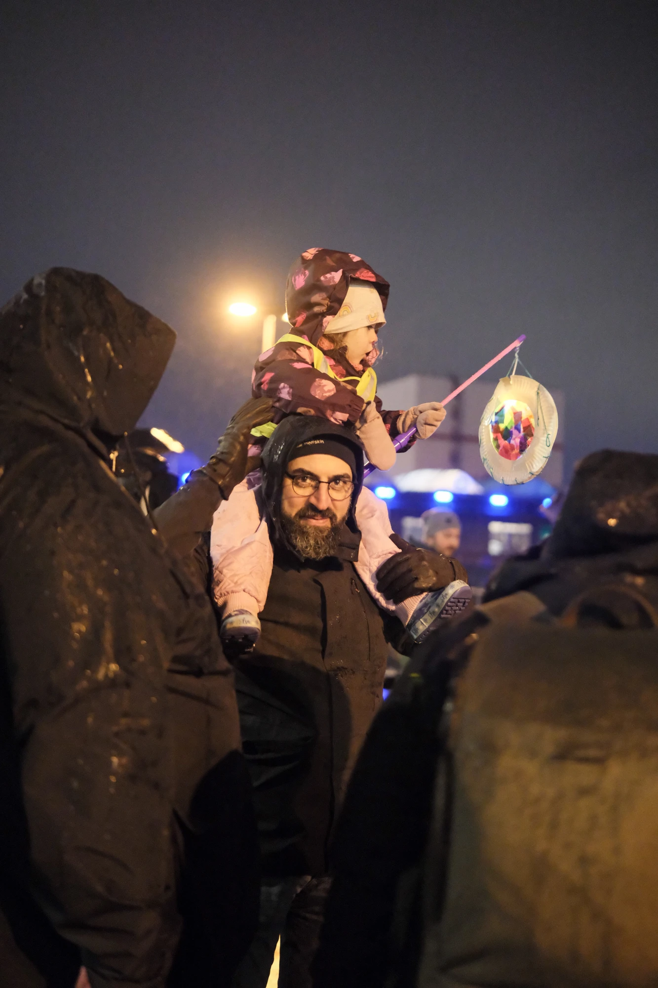 Kind mit bunter Laterne auf den Schultern eines Erwachsenen, umgeben von mehreren Personen in dunkler Kleidung bei Nacht.