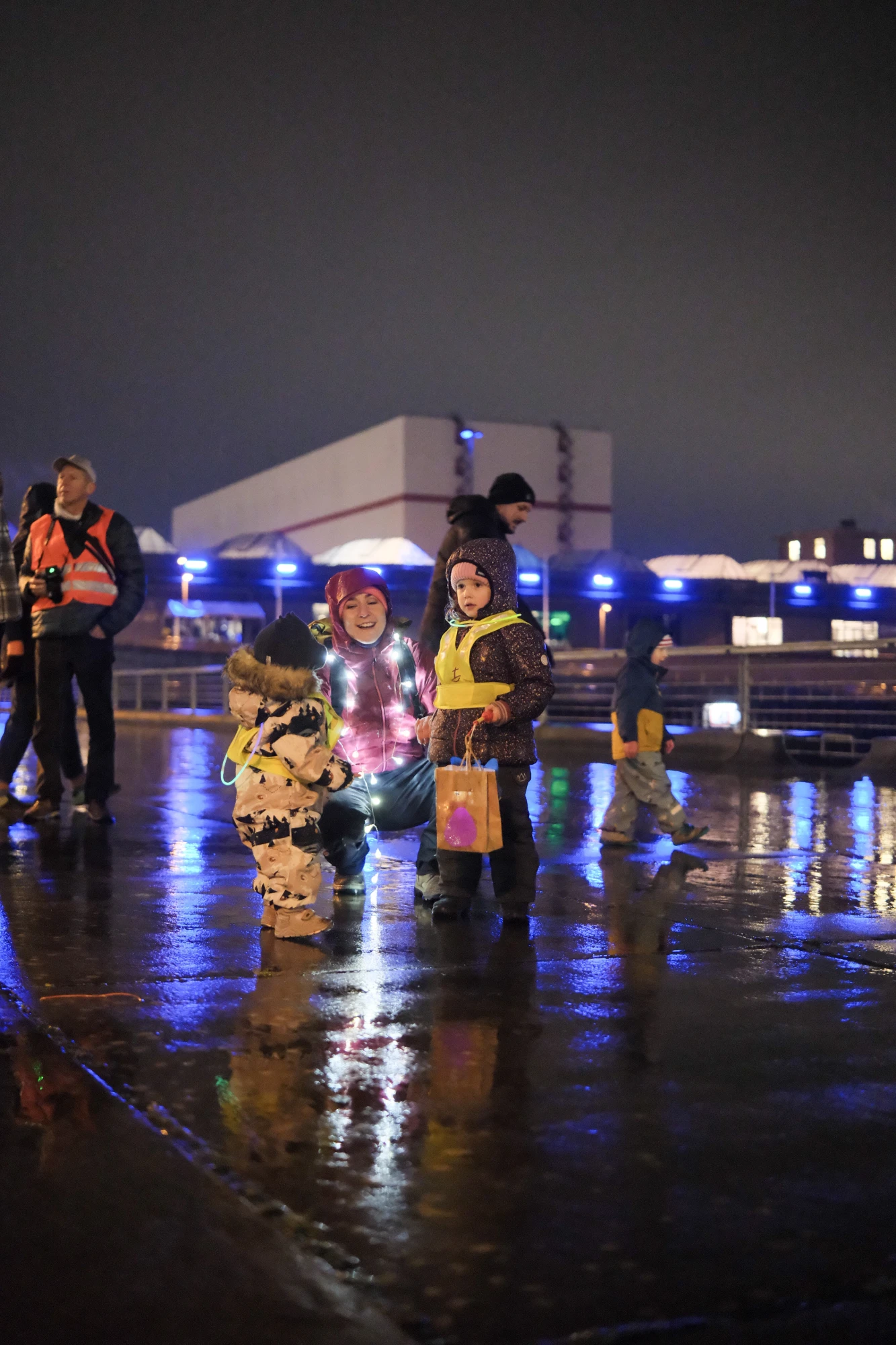 Mehrere Personen, darunter Kinder mit reflektierenden Warnwesten, stehen auf nassem Boden bei Nacht vor einem beleuchteten Gebäude.