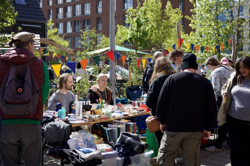 1.	Menschen stehen und sitzen an mehreren Tischen im Freien, die mit verschiedenen Gegenständen wie Kleidung, Geschirr, Büchern und Dekorationsartikeln belegt sind. Bunte Wimpelgirlanden hängen über den Tischen. Im Hintergrund sind Bäume, Sträucher und ein mehrstöckiges Backsteingebäude zu sehen. Die Szene spielt bei Tageslicht.