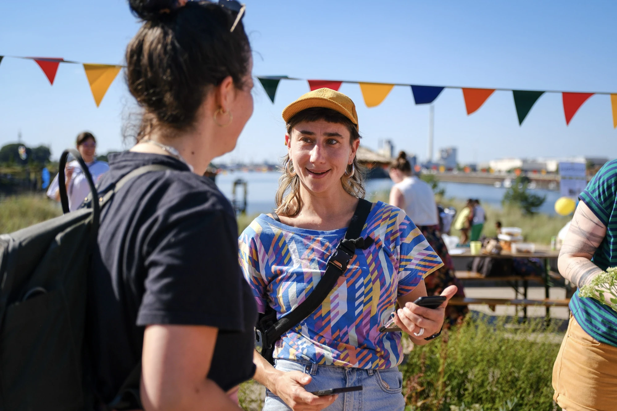 Zwei Frauen unterhalten sich im Freien bei sonnigem Wetter, eine trägt ein bunt gemustertes T-Shirt und eine gelbe Kappe, die andere ein schwarzes T-Shirt und einen Rucksack